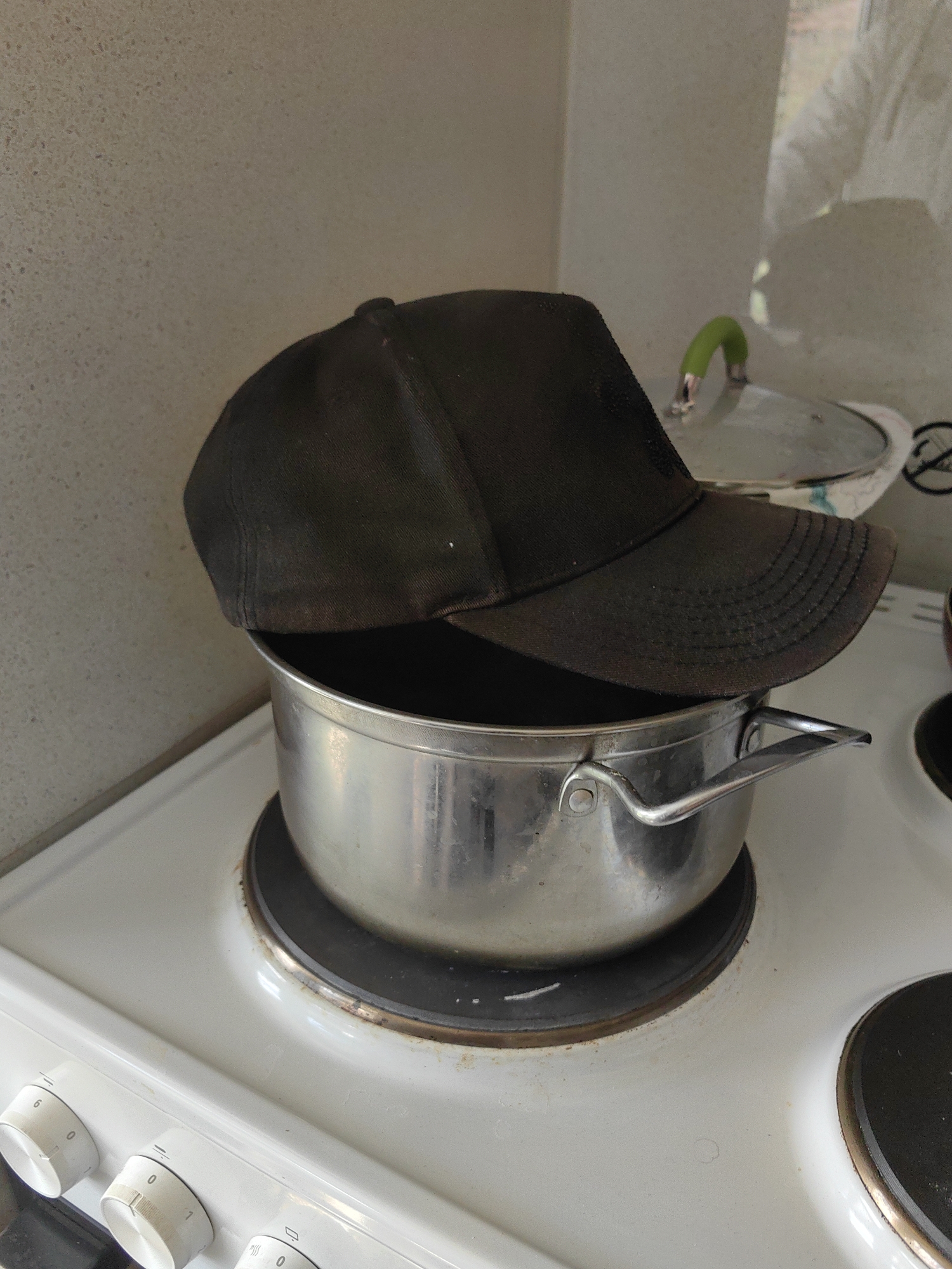 A black baseball cap sits atop a stainless steel cooking pot on a white electric stove. The stove has several control knobs on the front. Another cooking pot with a glass lid is visible in the background. The background includes a light-colored wall and a partially visible window. The location is inferred to be Hirevo, Bulgaria, based on the user request, though this information is not visually verifiable.