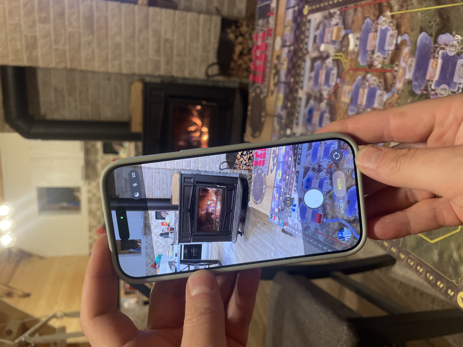 The scene is an indoor environment in Palež, Montenegro. In the foreground, two human hands hold a light-colored smartphone, encased in a pale green protective cover, vertically. The phone's screen displays a live camera feed showing a cast-iron wood-burning stove with a visible fire and a surrounding brick-patterned wall. To the left of the stove on the screen, a modern kitchen area with white cabinets and a stainless steel appliance is visible. On the bottom part of the phone screen, camera interface elements include a white circular shutter button, text labels "ВИДЕО" (Video) and "ФОТО" (Photo) in Cyrillic, and numerical zoom options (0.5x, 1x, 1.8x). An augmented reality (AR) overlay or digital representation of a board game, featuring purple and red structures on a map-like grid, is visible over the floor on the phone screen.
In the background, outside the phone's view, another black cast-iron wood-burning stove with a burning fire is present against a similar brick-patterned wall. To the right of this background stove, a black metal basket contains a stack of cut firewood. A large board game, identical in style to the overlay seen on the phone, is spread out on a dark surface in the lower right, featuring numerous purple, pink, and red game pieces. The room features a light-colored wooden ceiling with exposed beams and a modern light fixture with three spherical bulbs. A dark brown chair or stool is partially visible in the lower left.