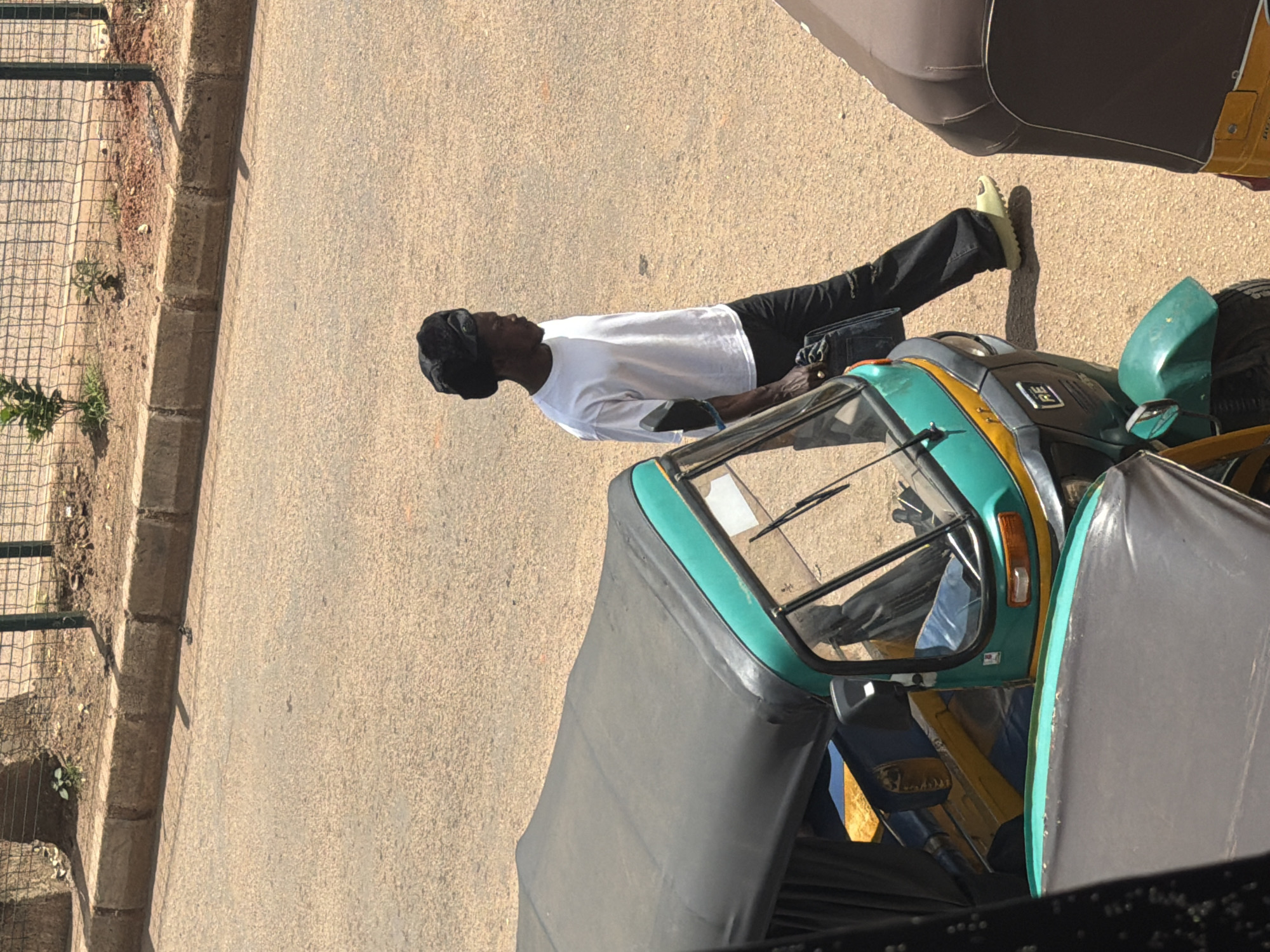 In the city of Bukuru, Nigeria, a Black man wearing a white t-shirt, black pants, and a black headscarf walks across a paved road. He is carrying a dark bag in his right hand. To his left, a green and yellow auto-rickshaw is parked on the road. To his right, another auto-rickshaw, brown and yellow, is partially visible. The scene is illuminated by sunlight, casting shadows on the road and the side of a bordering sidewalk. The sidewalk is made of stone and concrete, with a wire fence and some vegetation behind it.