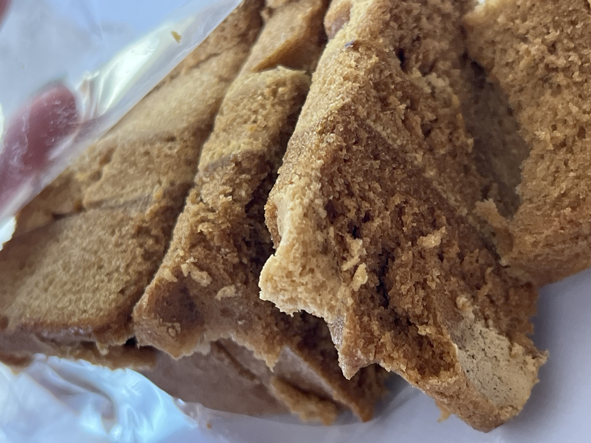 The image is a close-up, top-down view of several slices of brown bread, possibly cake or a dense loaf, stacked and contained within a clear plastic bag. The bread has a porous, slightly crumbly texture and a light brown, golden hue. The edges of the slices are uneven, suggesting they have been torn or roughly cut. Some slices are layered over each other, creating depth and shadow. A portion of the plastic bag is visible in the upper right corner, reflecting light and obscuring some of the bread. The background is a plain, light color, likely a surface the bag is resting on. The image does not contain any specific geographical markers, people, or actions, and therefore cannot be placed in Krasnyye Barrikady, Russia.