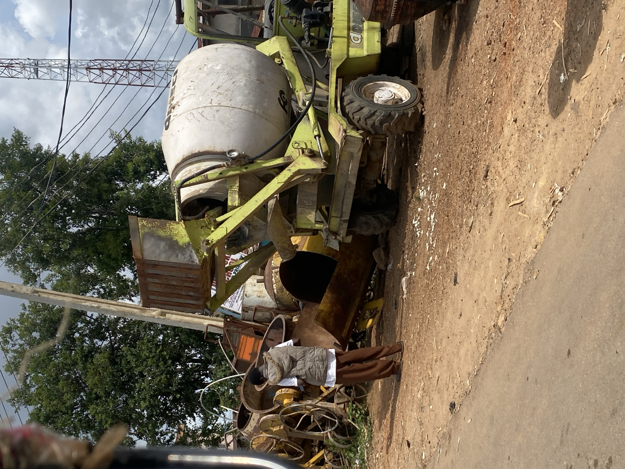 The image depicts an outdoor scene in Rafin Par, Nigeria, likely a construction site or scrapyard. In the foreground, a cement mixer truck with a light green chassis and a large white drum is parked on a dirt road. The truck appears weathered. To the left of the truck, a person wearing a light brown vest over a white shirt and brown pants stands facing away from the camera, looking at several old, rusty construction machines, including what appear to be old cement mixers with large wheels. The ground is covered in dirt, gravel, and some debris. In the background, lush green trees and a tall, red and white telecommunications tower are visible under a cloudy sky. Electrical wires crisscross the scene. The overall impression is one of industrial activity or storage of used heavy machinery in a somewhat unkempt environment.