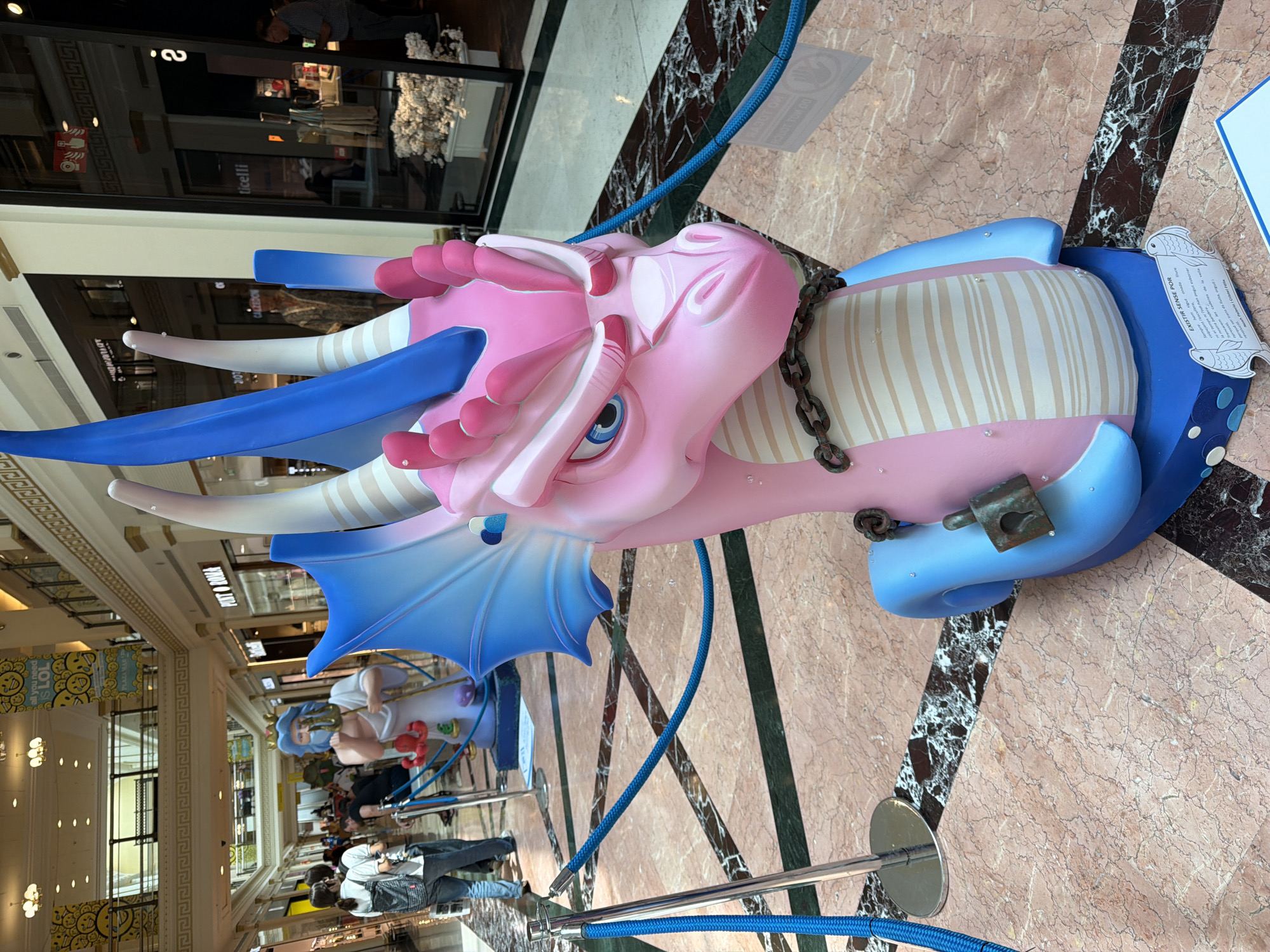 In the center foreground of the image is a large, pink and blue dragon sculpture. The dragon's head is facing right, with its mouth slightly open. It has blue wings, a long blue horn, and pink spikes on its head. A metal chain is wrapped around its neck, and a padlock is attached to the chain on the right side of its neck. The sculpture is standing on a blue base with white circles and two fish cutouts on the front. A small white sign with text is attached to the base. The dragon is in a mall with a marble floor. In the background, several people are walking and browsing in the mall. There are storefronts on either side of the mall corridor. Yellow banners with smiley faces are hanging from the ceiling. The scene appears to be a public indoor space, likely a shopping mall.