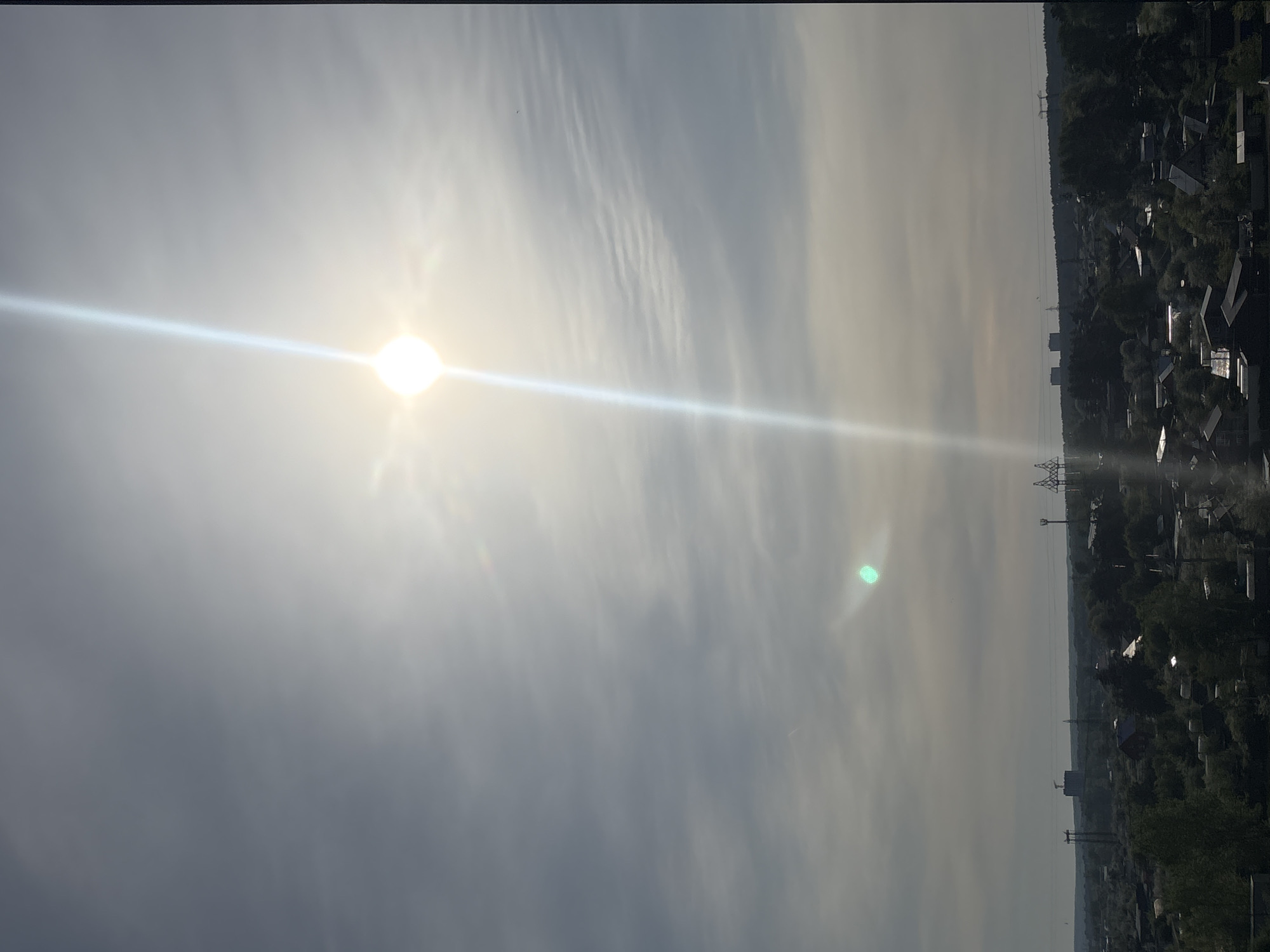 The image, taken in what appears to be Chaikovsky, Russia, depicts a hazy sky with the sun prominently visible and a lens flare extending across the frame. Below the horizon line sits a residential area with buildings interspersed among trees, seemingly viewed from an elevated vantage point. Transmission towers are interspersed among the structures. The atmosphere appears hazy.