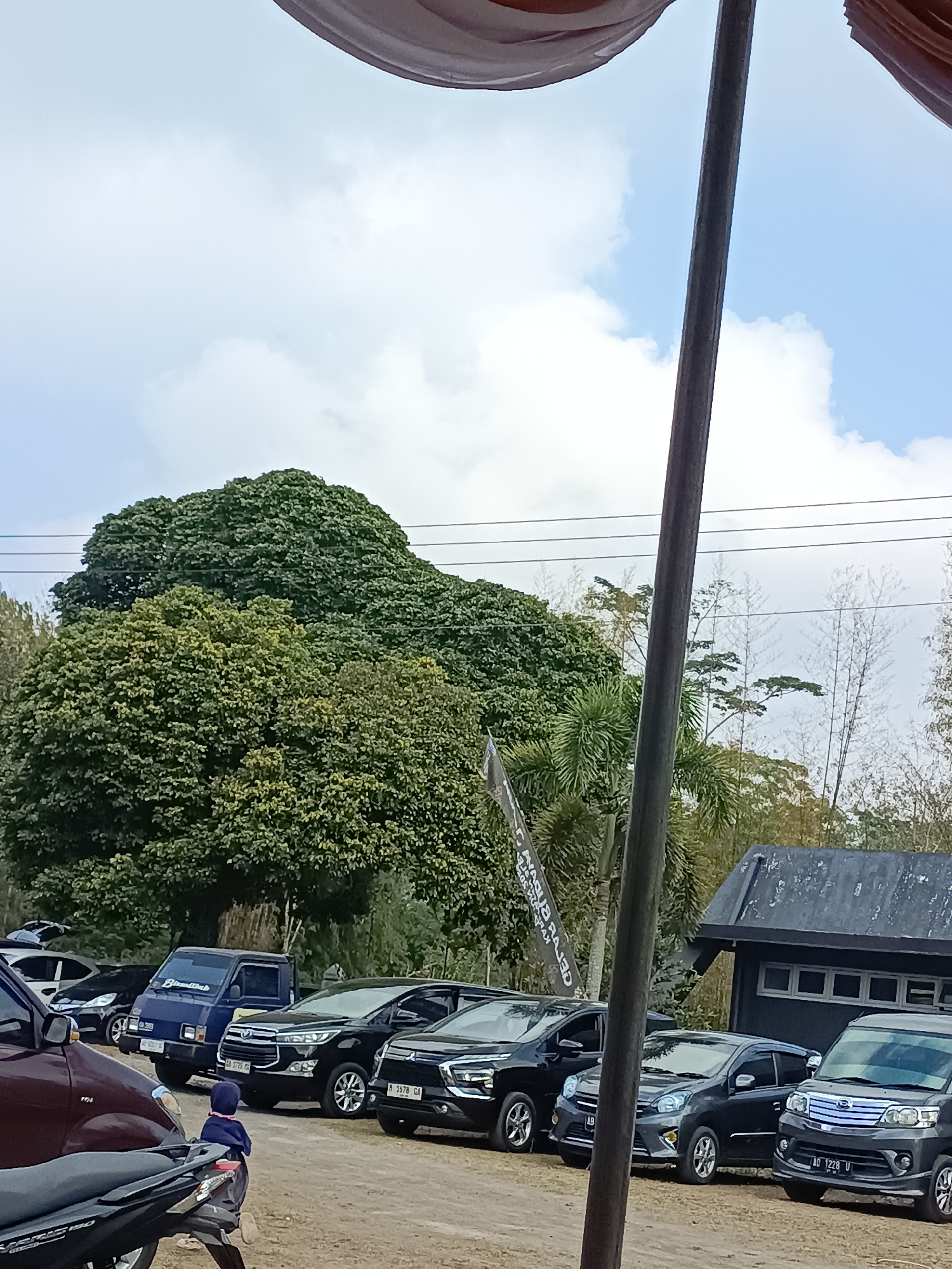 A low-angle, medium shot captures a parking lot filled with vehicles under a partly cloudy sky. In the foreground, the edge of a dark motorcycle is visible, with a maroon car parked to its left. A child wearing a blue hoodie and a dark blue shirt is seated on the rear of the maroon car.
Behind the maroon car, several cars are parked in a row: a black SUV, a black sedan, a dark gray small hatchback, and a dark gray minivan. Further back, a blue pickup truck is parked. A large, dark green, rounded tree dominates the middle ground, with more trees and bamboo visible to the right.
A rustic, dark-colored building with visible windows is situated behind the parked cars on the right side of the frame. A flagpole with a banner hangs to the left of the building. Power lines stretch horizontally across the upper portion of the image, above the trees. The ground appears to be unpaved, likely dirt or gravel. The scene is likely outdoors, possibly in a rural or semi-rural area, given the presence of trees and the unpaved lot. Based on the text on the banner, which reads "PUSKESMAS HARGO</>BINANGUN", the location is likely the Puskesmas (community health center) in Hargobinangun, Sleman Regency, Yogyakarta Special Region, Indonesia.