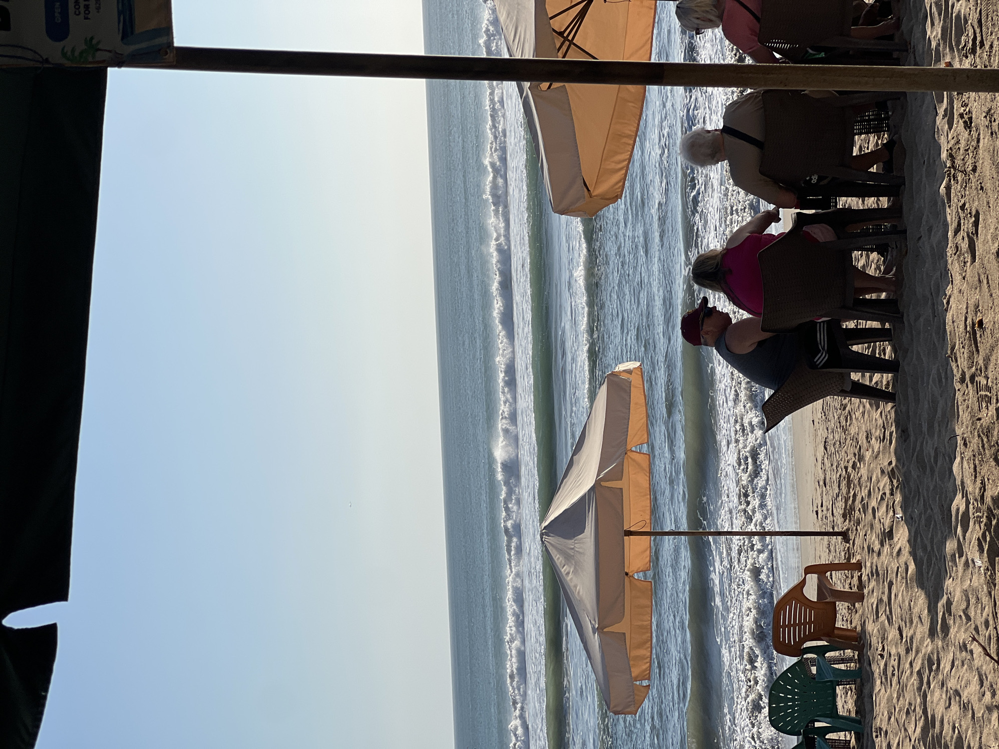 An outdoor cafe scene on the sandy beach in Kuta, Indonesia, is depicted under a clear blue sky. The ocean, with its characteristic turquoise hue, features gentle waves rolling towards the shore. Two large, light-colored umbrellas provide shade for patrons seated at tables. In the foreground, several plastic chairs are scattered on the sand, including a green chair and an orange chair positioned near the water's edge.
A group of four individuals are seated at a table under one of the umbrellas. A man wearing a dark tank top and a red cap is seated on the left, facing away from the camera. Beside him is a woman in a bright pink top. To their right, a man with white hair, wearing a light brown shirt, is seated, facing the ocean. A woman in a light pink top is seated on the far right, also facing the ocean. They appear to be engaged in conversation or enjoying the view.
The scene is characteristic of a beachfront establishment in Kuta, a popular tourist destination known for its beaches and vibrant atmosphere. The overall impression is one of relaxation and enjoyment in a tropical setting.