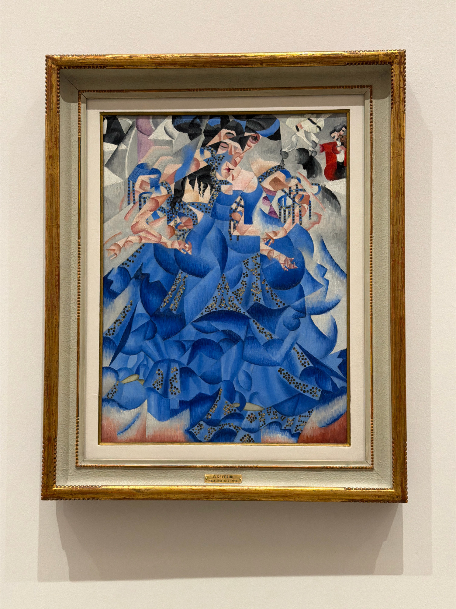 The image shows a painting in a golden frame, displayed on a white wall. The painting is an abstract depiction of figures in vibrant blue and other colors, with geometric shapes and fragmented forms. In the lower part of the frame, a small plaque is visible. A shadow is cast on the wall to the left of the painting. The scene appears to be in a museum or art gallery. The plaque at the bottom of the painting reads "G. SEVERINI "DANSE BLEU" 1912". This suggests the painting is "Danse Bleue" by Gino Severini, created in 1912. Gino Severini was an Italian painter and a leading member of the Futurist movement. While the painting itself is the primary subject, its likely display in a museum, potentially in Milan, Italy, where Futurist art is often exhibited, provides context.
