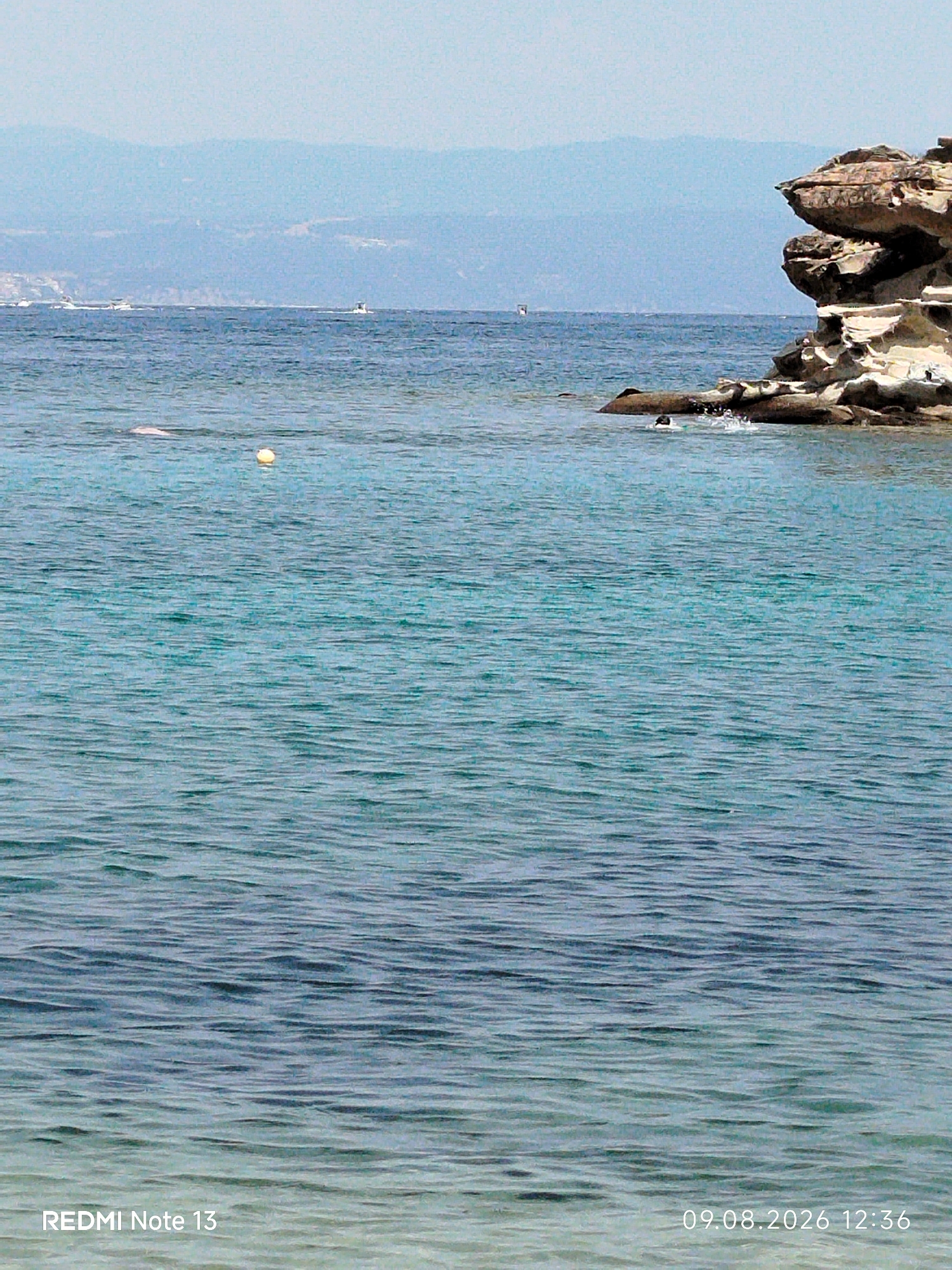 The image depicts a serene coastal scene in Sithonia, Greece, likely during daytime. The foreground is dominated by tranquil, shimmering blue-green water, characteristic of the Aegean Sea. Gentle ripples suggest a calm atmosphere. In the mid-ground, several boats are visible on the horizon, hinting at leisure activities or transportation. A yellow buoy floats serenely on the water, and a person is partially submerged near a rocky outcropping, possibly swimming or engaged in water sports, with a splash suggesting movement. The rocky formation, prominent on the right side of the image, is sculpted by natural erosion, showcasing layers of sediment and a rugged texture against the clear sky. In the background, distant mountains create a soft, hazy silhouette against a pale blue sky. The overall impression is one of natural beauty and peaceful outdoor recreation.