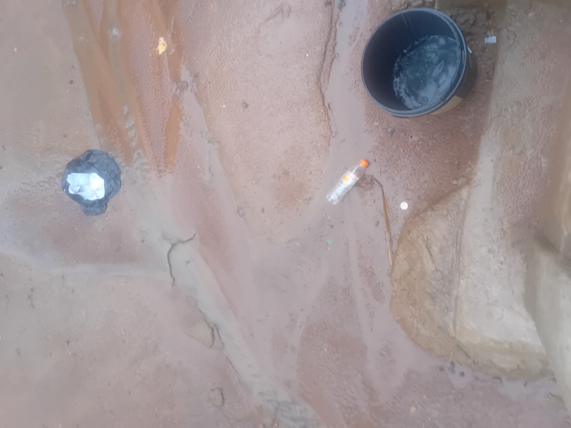In Jauro Yinu, Nigeria, a scene of a muddy ground is depicted. A black bucket filled with water sits to the right of the frame. Nearby, a plastic bottle with an orange cap lies on its side. Further up, a small pile of debris, possibly including plastic, is visible. The ground itself is uneven, with visible water channels and patches of dry mud. The overall environment appears to be an outdoor area, possibly a poorly maintained pathway or drainage area.