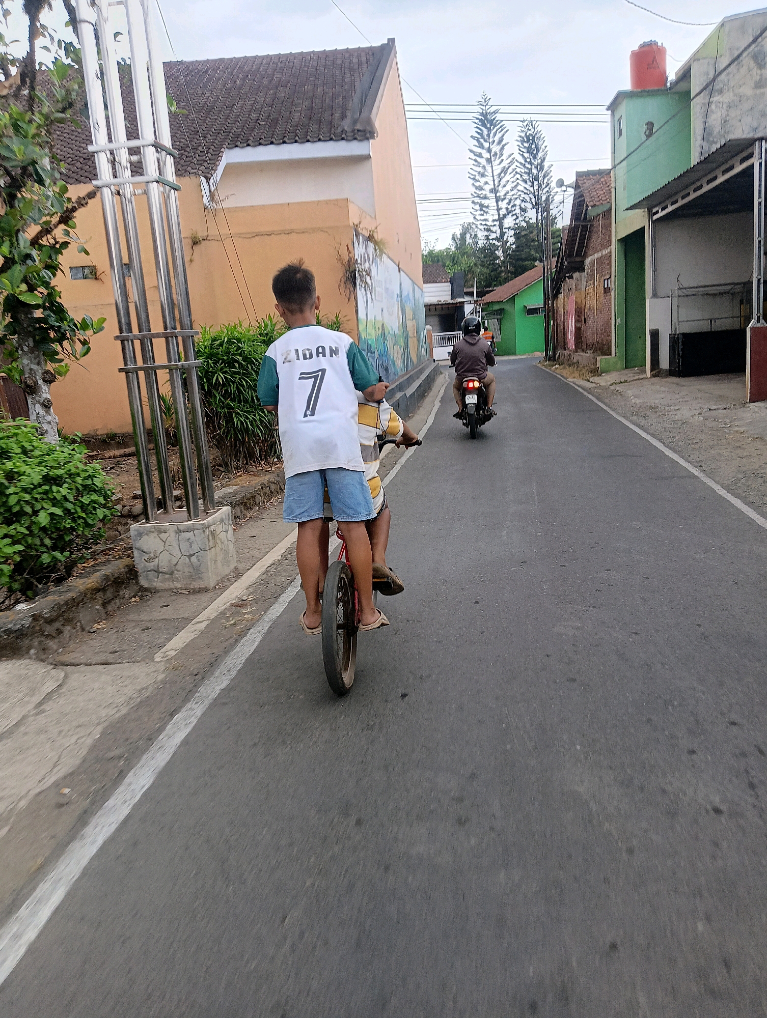 In Sokaraja, Indonesia, a boy wearing a white jersey with the name "ZIDAN" and the number "7" on the back, along with blue denim shorts and flip-flops, rides a bicycle on a paved road. He is positioned in the foreground, with his back to the camera. The bicycle has a single red frame and a thin black tire. Another person is riding a motorcycle in the background, facing away from the camera. The road is bordered by buildings on both sides, with some trees and lush green vegetation visible. The buildings are a mix of colors and architectural styles, with one notable orange building and a green building with an orange water tank on its roof. The sky is overcast. The scene appears to be a residential street in a moderately developed area.