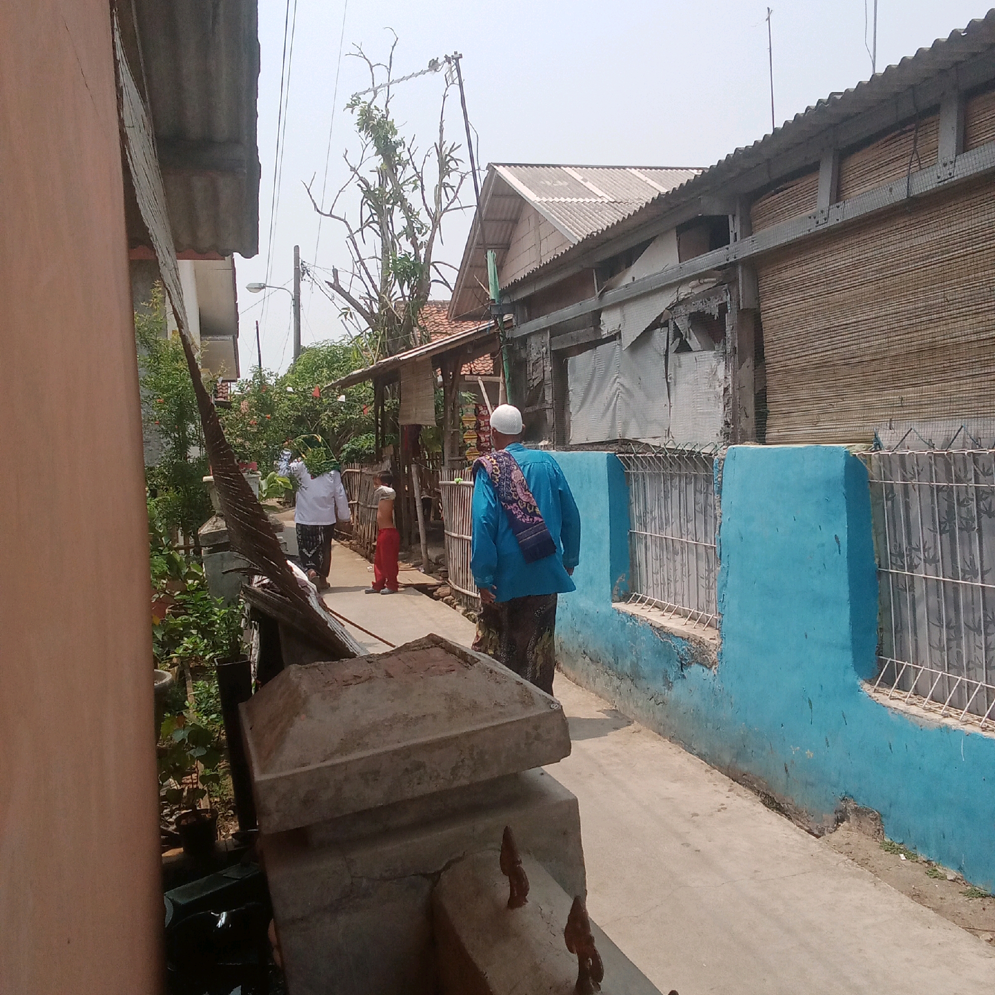 The image depicts a narrow street in a residential area, possibly in Karawang, Indonesia. The scene shows three individuals walking away from the viewer. The person in the foreground on the right is wearing a bright blue long-sleeved shirt, a patterned sarong, and a white cap. They have a patterned scarf draped over their shoulders. To their left and slightly ahead, another person is visible from behind, wearing a white long-sleeved shirt and a sarong, with a green and yellow headscarf. Further ahead and to the left is a child wearing red pants.
The street is paved with concrete and is bordered on the right by a vibrant blue wall with a metal fence. Behind the fence, a building with a corrugated metal roof and bamboo blinds is visible. On the left side of the street, there are more buildings with corrugated roofs, as well as lush green foliage and trees. A tall, thin utility pole with wires runs vertically in the background, partially obscured by trees. In the foreground on the left, a part of a brown-colored structure and a gray concrete object are visible, suggesting a building or a raised platform. The overall impression is of a typical urban or suburban street scene in a tropical climate, likely in Indonesia, given the attire of the individuals.