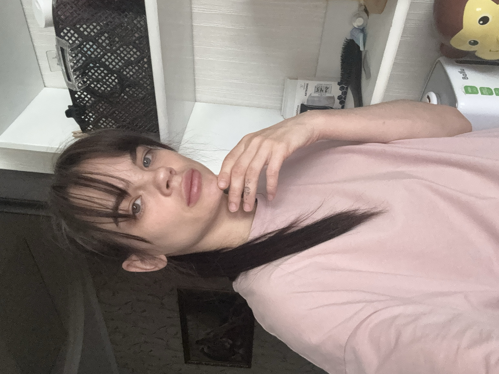 The media file presents a self-portrait of a young woman, captured indoors, likely in Rostov-on-Don, Russia. The image is vertically oriented (rotated 90 degrees clockwise from a standard upright view), showing the woman from a slightly low angle.
The subject is a young woman with dark hair and bangs, framing a face with light-colored eyes and a neutral, slightly pouting expression as she looks directly at the camera. She is wearing a light pink t-shirt. Her right hand, adorned with a faint tattoo on her fingers, is gently placed near her chin and mouth.
The background suggests a domestic setting. White shelving units are visible, holding various household items. On the upper left, a dark, grid-patterned basket is present. On the upper right shelves, a small, yellow and brown soft toy, resembling a hedgehog, is visible. Below it, a white and grey appliance, possibly a small humidifier or heater, is branded with "Ballu" and features green control buttons. Adjacent to the appliance, white packaging with partially visible text "AKTRONIK", "OZO", and "245" can be seen. A dark hairbrush is also on this shelf. The wall below the shelves is covered with a dark, patterned wallpaper, on which a dark-framed picture or mirror hangs. The lighting suggests an indoor setting, likely during the daytime. The overall impression is a candid, personal moment captured within a home environment.
