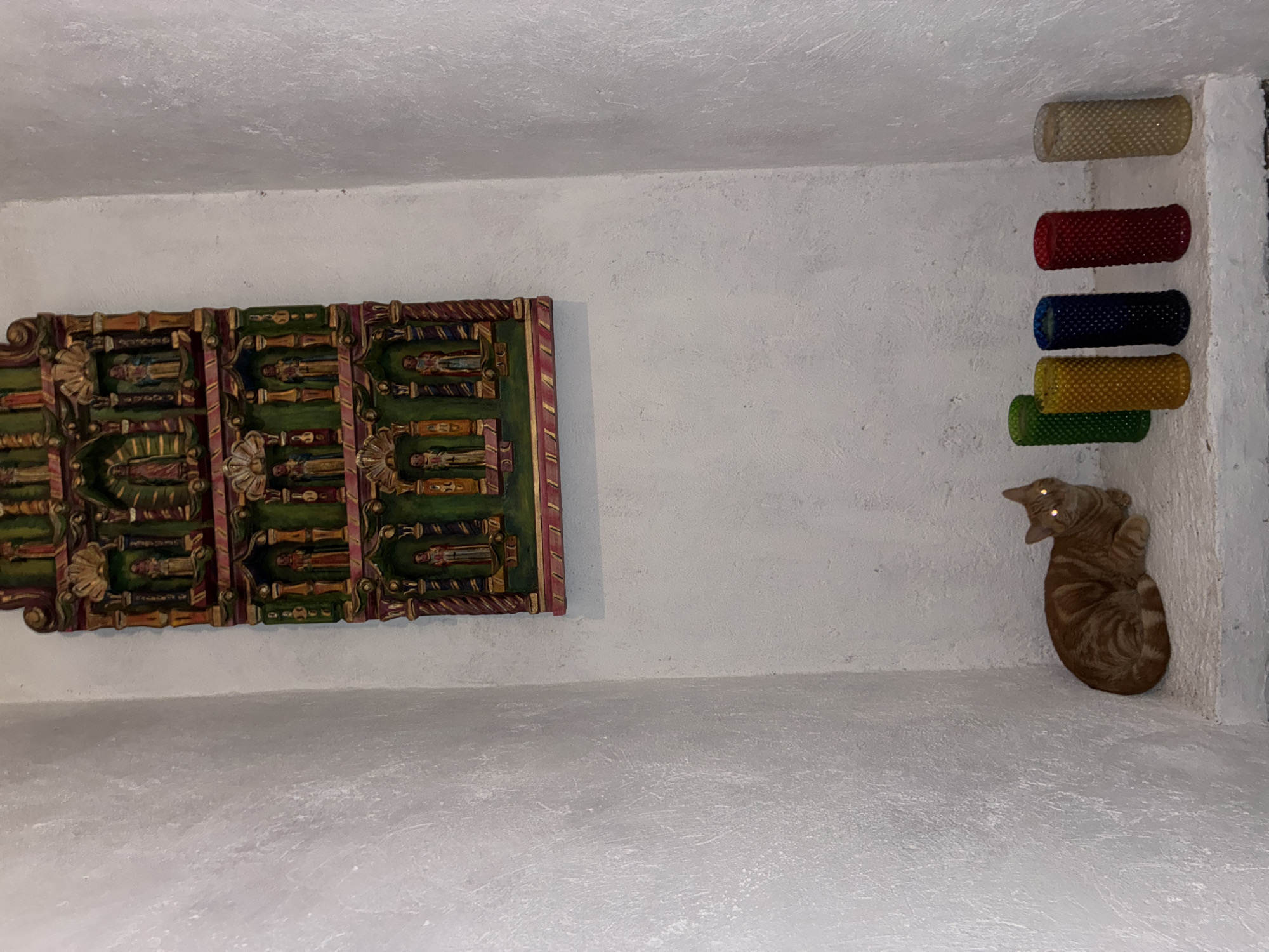 A low-angle shot depicts an interior corner of a room. On the left wall hangs a decorative wooden piece featuring repetitive figures in a vertically oriented rectangle, adorned with gold, green, and red-brown tones. To the right, cylindrical objects colored white, red, blue, yellow, and green are attached to the wall, arranged vertically. A cat with tabby-striped fur is curled up at the intersection of the walls and floor. The walls are textured, and the location may be Cañada de Madero, Mexico.