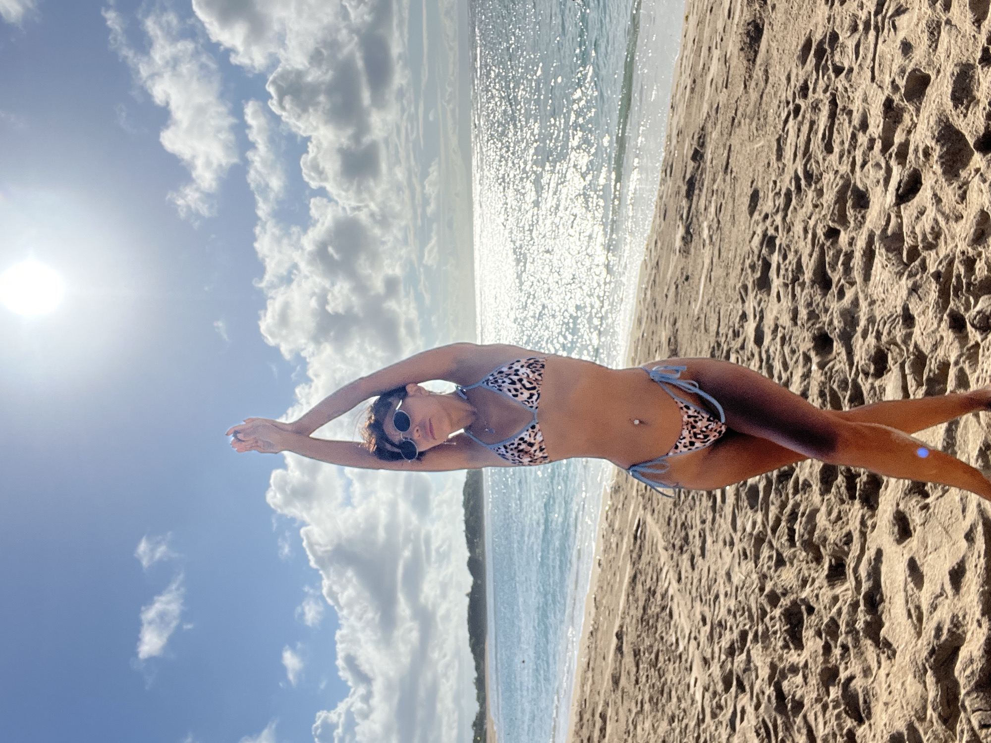 The photograph depicts a woman lying on a sandy beach. She is wearing a bikini with a leopard print and blue ties, and sunglasses. Her arms are raised overhead. The scene includes a beach, the ocean, and a sky with clouds and the sun. The location is Kuta Selatan, Indonesia.