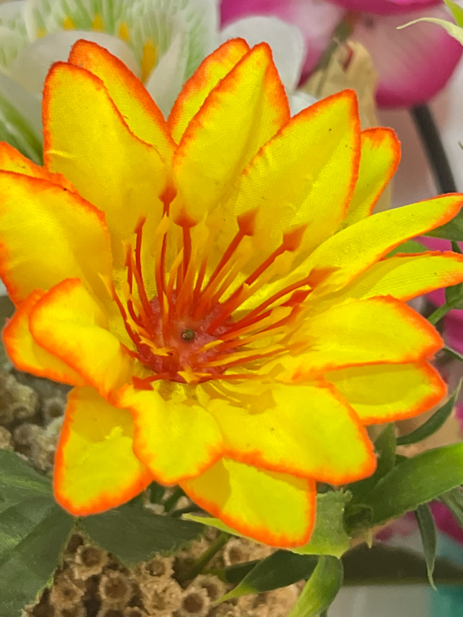 A close-up, eye-level shot focuses on a vibrant, artificial yellow flower with orange-tipped petals. The flower's center is a cluster of red stamens. In the background, out-of-focus flowers in shades of white and pink are visible. The scene is indoors, possibly a home or a shop in Denpasar, Indonesia, where such decorative flowers are common.