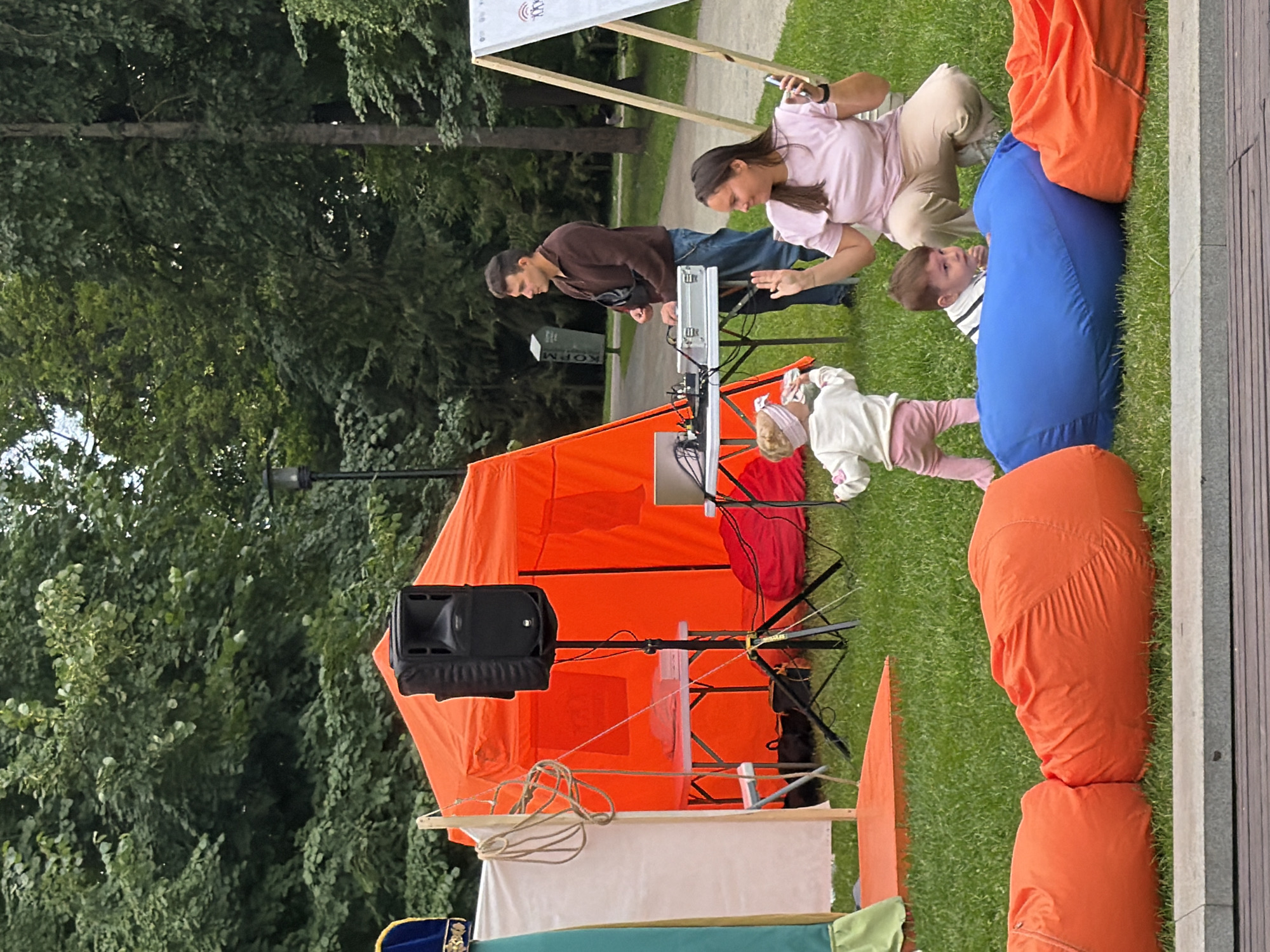 The image depicts an outdoor gathering with several people and objects set against a backdrop of lush green trees. In the foreground, a bright orange tent is set up, with a black speaker mounted on a stand next to it. Various ropes and fabric elements are draped around the tent, suggesting a festive or event-like atmosphere.
Several individuals are present, including a man and a woman interacting with a DJ setup that includes a silver console. A young child is visible near the DJ setup, and another child is seen further back, dressed in pink and white. The woman is wearing a light pink top and beige pants, while the man is in a brown jacket and jeans.
In the background, the scene transitions to a paved path, with more trees providing a natural setting. The lighting suggests it is daytime, possibly late afternoon, with diffused sunlight filtering through the foliage. The overall mood appears relaxed and casual.
A partially visible sign with text can be seen near the top of the image, but the content is not clearly legible. The presence of the tent, DJ equipment, and multiple people suggests an outdoor event or performance is taking place. The setting appears to be a park or open public space.
