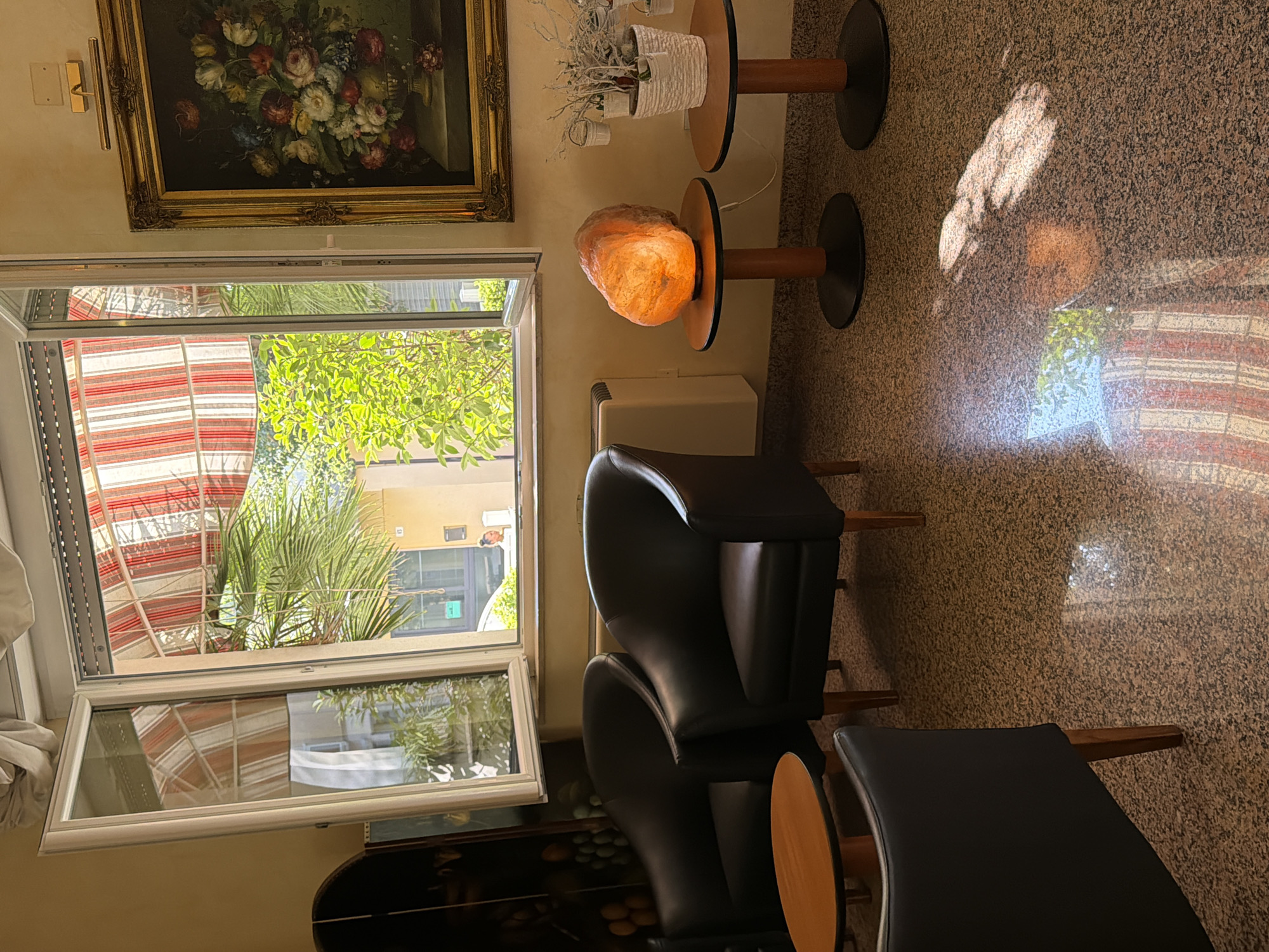 The image depicts an indoor scene, likely a hotel lobby or lounge area in Gatteo, Italy. The room features dark leather armchairs, two round wooden side tables, and a speckled granite floor. Sunlight streams in through an open window, illuminating the space and casting reflections on the floor. Through the window, an outdoor scene with lush greenery and buildings can be seen, along with a red and white striped awning. On one of the side tables, a large, glowing orange salt lamp is prominently displayed, while the other table holds a small potted plant with white decorative elements. A framed painting of a floral still life hangs on the wall to the right, illuminated by a small brass picture light. The overall atmosphere suggests a comfortable and well-lit waiting area.