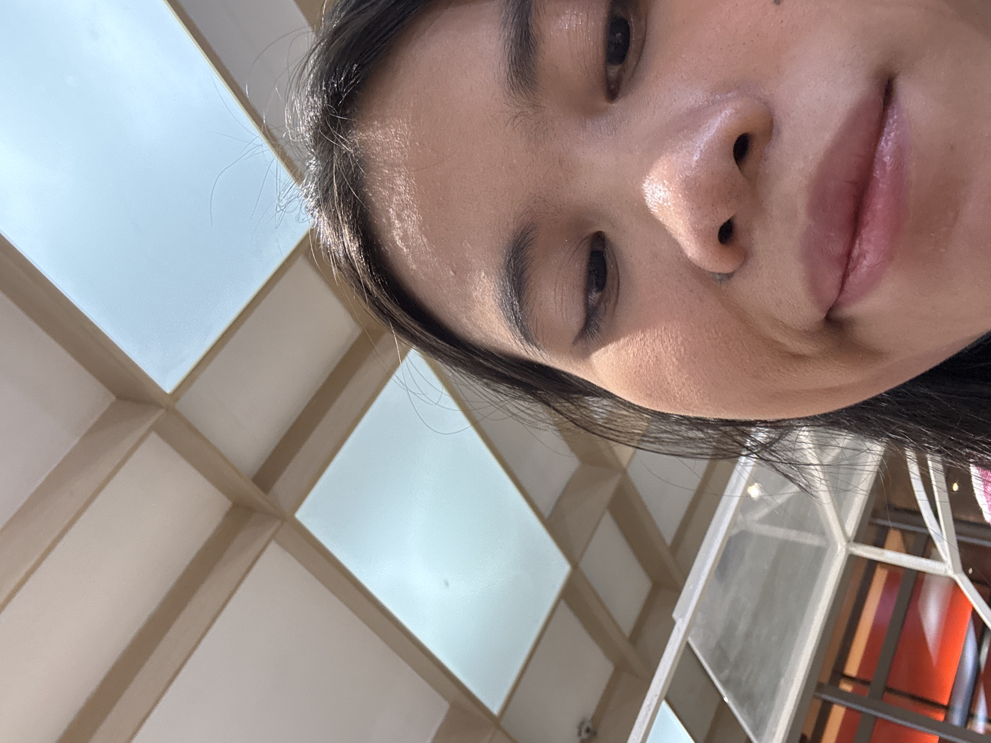 The image is a close-up, low-angle selfie of a person's face, with a ceiling structure and a glimpse of an interior space in the background. The person's eyes are looking downwards, and their lips are slightly parted in a subtle smile. The ceiling is composed of rectangular panels and beams, with sections appearing to be translucent or made of frosted glass, letting in natural light. The background interior shows a modern architectural design with red and orange elements visible through a glass partition. The image quality is adequate for identification purposes. No specific location in Central Jakarta, Indonesia, can be determined from the visual information presented in the image.