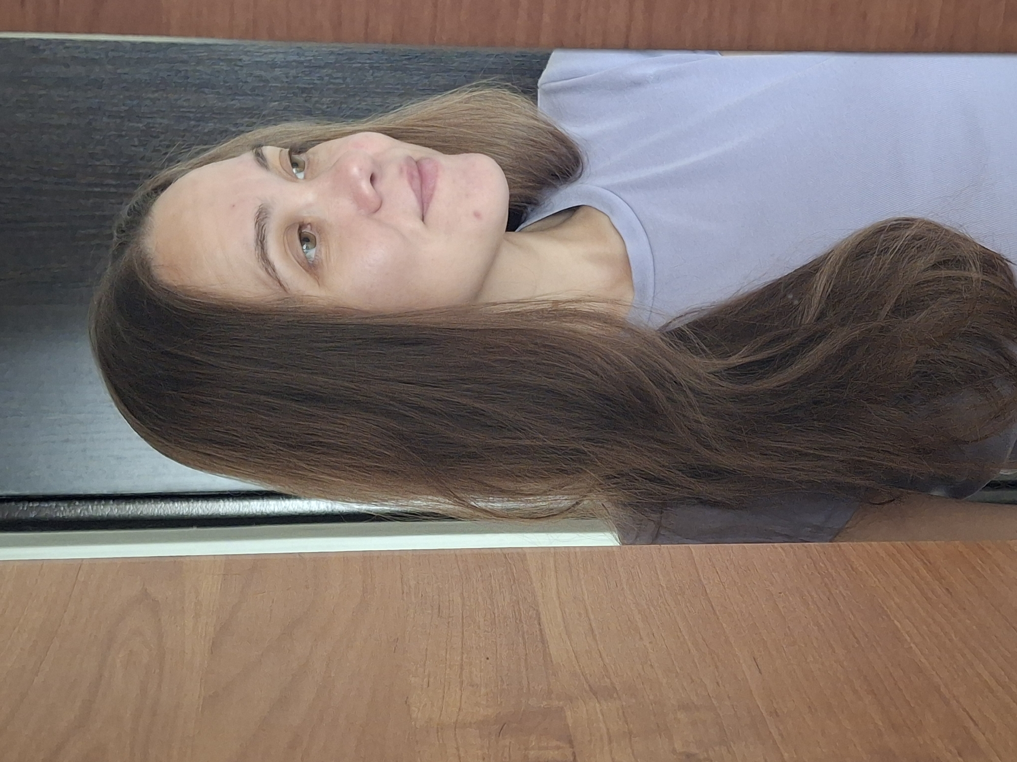 A lone adult female is depicted in a mid-shot portrait, captured in a right-facing profile. Her head is turned to the right, and her gaze is directed slightly upwards and to the right, beyond the frame. A subtle, closed-mouth smile is visible on her face. She has long, wavy, dark brown hair that cascades over her right shoulder and partially down her chest. Her complexion appears fair with natural skin tones, and minor moles or blemishes are visible on her forehead and chin. Her eyes are light brown or hazel, and her eyebrows are well-defined and natural. She is wearing a plain, light lavender or periwinkle blue crew-neck t-shirt. The scene appears to be indoors. To the immediate left, a light brown wooden surface with prominent vertical grain occupies approximately one-third of the frame. A thin white vertical trim separates this wood from a darker, textured vertical surface which appears dark grey or charcoal in color, spanning the middle ground behind the subject. This darker surface has subtle vertical lines. A partial glimpse of another wooden surface, similar to the one on the left, is visible at the far right edge of the frame. Soft, ambient light illuminates the subject from the front-left, creating subtle shadows and highlights. The image itself provides no discernible visual cues or metadata to indicate a specific geographic location such as the city of Chaikovsky, Russia.