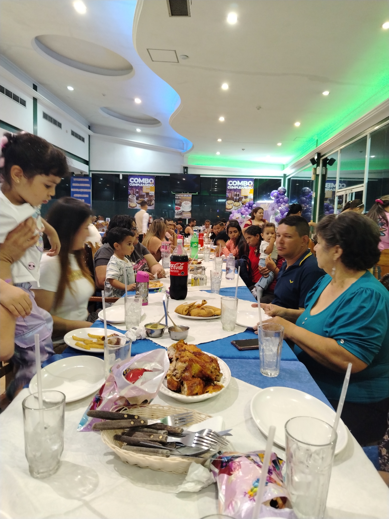 The image shows a family gathering at a restaurant, likely a birthday celebration, in Tinaquillo, Venezuela. A long table covered with a white and blue tablecloth is set for a meal with plates of food like fried chicken and fries, and a bottle of Coca-Cola. A basket of silverware is on the table, along with glasses and straws. Several people are seated at the table, including women, men, and children. Some are interacting with each other, while others are focused on the food. Two signs in the background advertising a birthday "combo" meal with a photo of a cake. Purple balloons are seen near the glass windows. The ceiling has recessed lighting with a wave-like design illuminated by blue and green light.