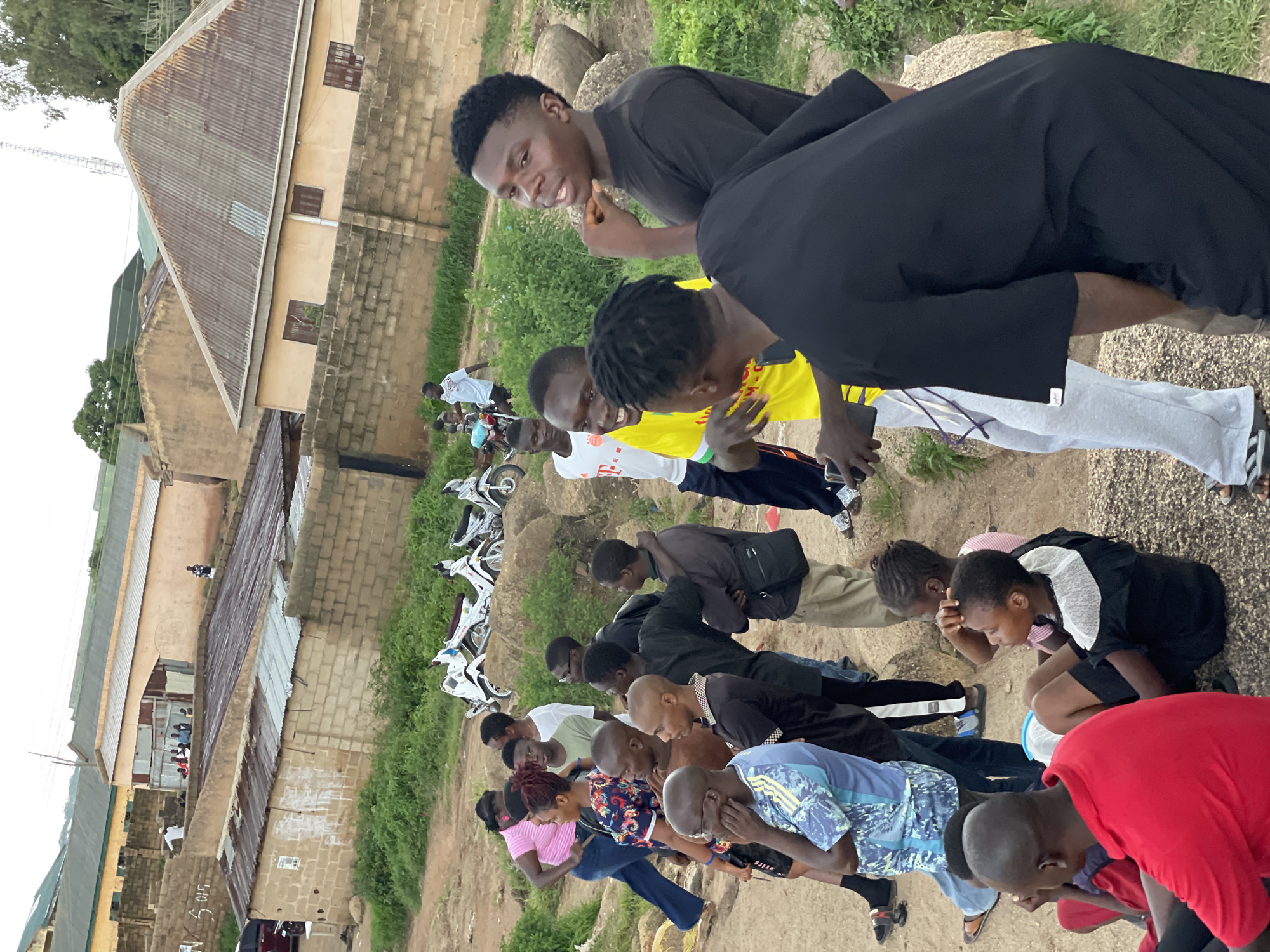 A group of young people, appearing to be mostly male, are gathered outdoors in what seems to be a somewhat dilapidated urban or semi-urban setting, possibly in Jos, Nigeria. The scene is viewed from a slightly elevated, tilted perspective. In the background, there are buildings with corrugated iron roofs and brick walls, some showing signs of wear. Green vegetation and large rocks are also visible, suggesting an outdoor area with some natural elements. Several motorcycles are parked in the middle ground, indicating a common mode of transport in the area.
The individuals are engaged in an activity that looks like a communal prayer or a ritualistic gathering, with most of them kneeling or bowing their heads in unison. Their expressions are mostly subdued, suggesting a serious or contemplative mood. The clothing varies, with some wearing t-shirts, others more formal shirts, and some in traditional garments. The lighting suggests it might be daytime, possibly late afternoon, with diffused light and no harsh shadows.
One of the more prominent figures in the foreground is a young man in a bright red t-shirt, his head bowed. Other individuals in the group are also in similar prayerful postures. In the upper part of the image, a few individuals are standing or observing, including one person wearing a black t-shirt and another in a black robe-like garment. There are no clearly legible text inscriptions visible in the image. The overall impression is one of a community engaged in a spiritual practice in a modest environment.