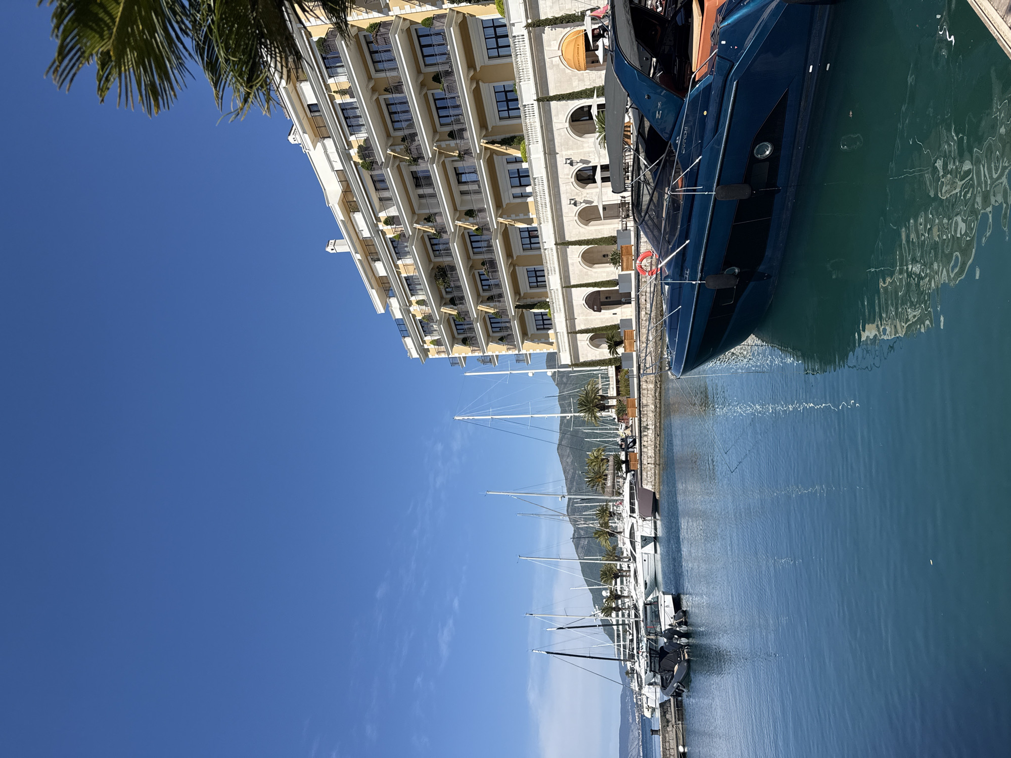 The image, oriented correctly, captures a vibrant marina scene under a clear blue sky in Tivat, Montenegro. In the upper left corner, the green fronds of a palm tree partially obscure the sky.
A grand, multi-story building dominates the left side of the frame, featuring a light yellow facade with numerous white-balustraded balconies and dark-paned windows. Its lower levels showcase arched entrances and windows, with a sign visible in one archway that appears to read "PROMENADE RISTORANTE."
Directly in front of this building, a marina is bustling with docked vessels. A sleek, dark blue or black motor yacht is prominent in the mid-ground, bearing the name "CRANCHI" on its hull and showcasing a tan interior. A red lifebuoy hangs from its side. Further along the marina, a line of white sailboats and smaller motor yachts are neatly moored, their masts reaching towards the sky.
The calm, clear turquoise water of the marina reflects the surrounding boats and the bright blue sky. In the background, across the water, majestic mountains rise under a largely cloudless sky, with only a few faint wisps visible. The scene is bathed in bright daylight, suggesting a sunny morning or early afternoon, contributing to an overall impression of luxury, leisure, and pleasant weather.