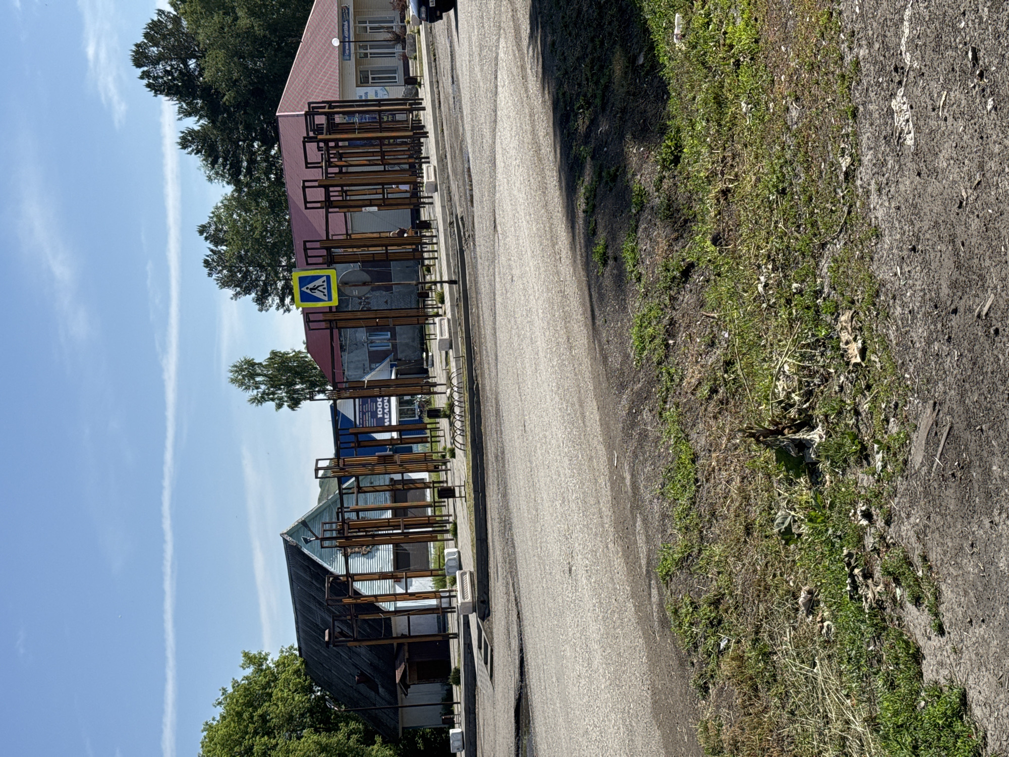 The image depicts a street scene in Soloneshnoye, Russia, under a clear blue sky with wispy white clouds. In the foreground, a rough asphalt road occupies the lower half of the frame, with patches of dry grass and dirt along its edge. Beyond the road, a sidewalk area features a distinctive metal structure with numerous vertical bars, suggesting it may be a decorative element or part of a playground. Several people are visible, with two individuals standing within the fenced area of the metal structure.
To the left of the structure, a building with a dark sloped roof and light-colored siding stands. To the right, a building with a red tiled roof and white exterior is visible, featuring multiple windows and a sign with text, some of which appears to be in Cyrillic. A yellow pedestrian crossing sign with a black symbol is prominently displayed near the metal structure. The overall environment suggests a public space, possibly a town square or park area, with surrounding buildings and a road for vehicular traffic.