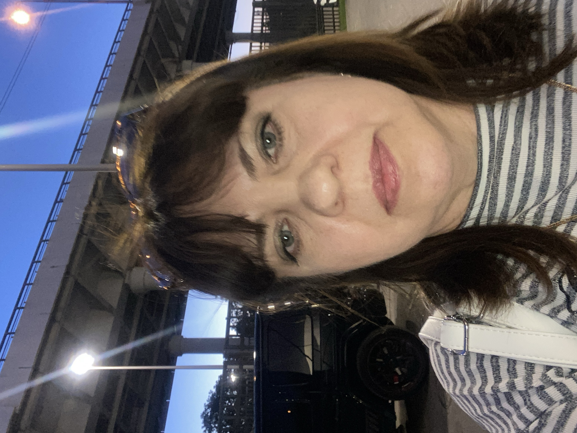 A middle-aged woman with dark brown hair and bangs takes a close-up selfie outdoors at twilight or night in an urban setting identified as Moscow, Russia. She wears a horizontally striped long-sleeved shirt (white and dark stripes) and a white shoulder bag with a silver buckle. Sunglasses are perched on top of her head. Her expression is neutral, and her eyes are light blue-green with subtle makeup. The background features a large concrete elevated roadway or bridge structure with support columns and a metal railing. Bright streetlights illuminate the scene; one on the left emits a strong white glare, and another on the upper right has an orange glow. To the left of the woman, the rear side of a dark-colored Mercedes-Benz G-Wagen SUV is partially visible, showing its distinctive wheel and a red brake caliper. Dark foliage is present in the distant left background.
