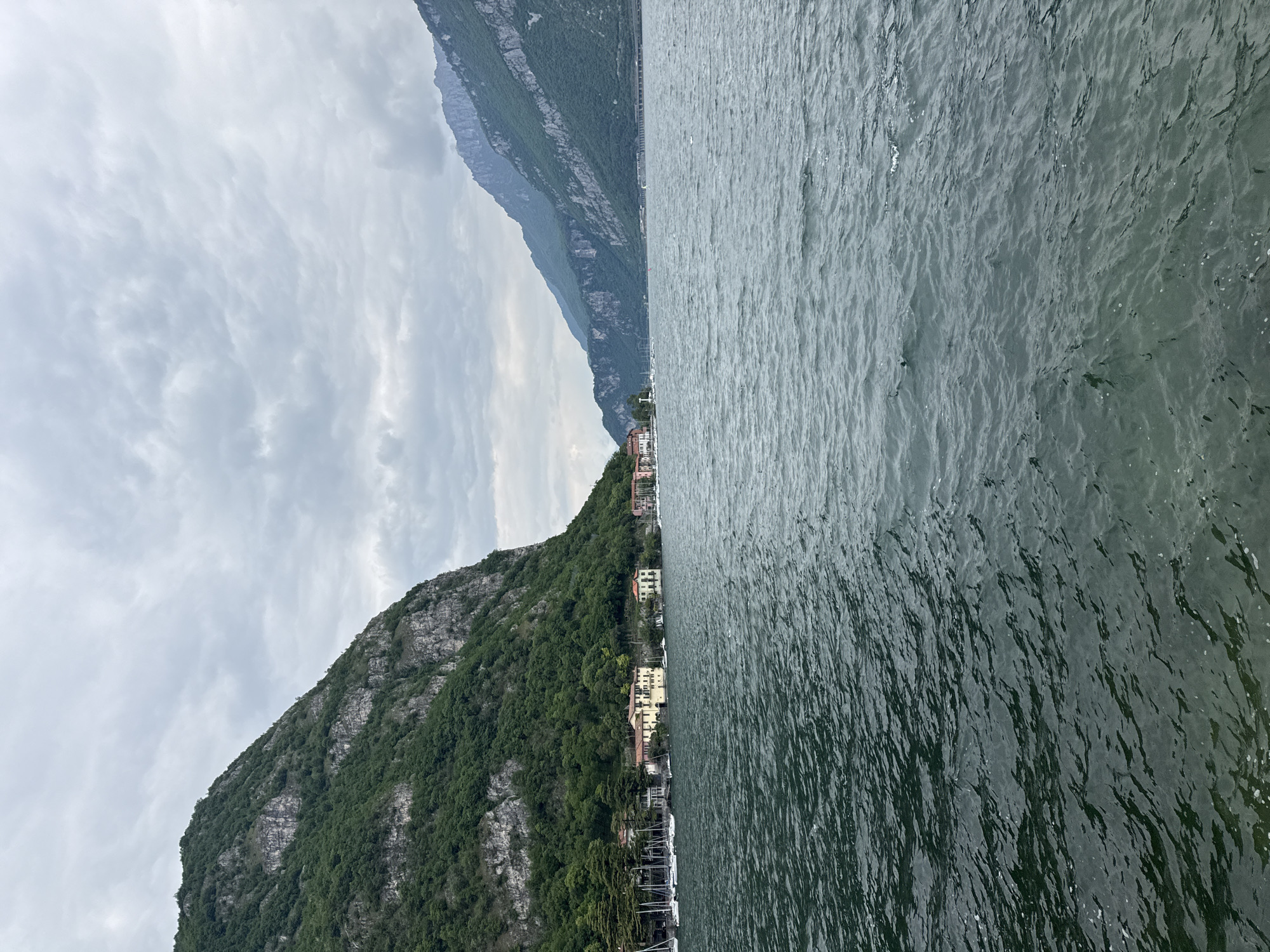 The image captures a serene landscape in Valmadrera, Italy, featuring a large lake bordered by steep mountains under an overcast sky. The right side of the frame is dominated by the expansive, dark green-blue water of the lake, its surface covered in numerous small ripples, reflecting the subdued light from above.
On the left, a towering mountain rises dramatically from the water's edge, its slopes densely covered with lush green trees and interspersed with rugged, grey rocky outcrops. Along the base of this mountain, a quaint settlement is visible, consisting of several buildings with light-colored facades—including shades of yellow, pink, and white—and traditional terracotta roofs. These structures appear to be houses or small lakeside establishments. In front of these buildings, along the shore, several boats, likely sailboats, are moored, their masts creating a pattern against the darker water.
The sky above is completely overcast, filled with soft, grey clouds that diffuse the daylight, preventing harsh shadows and contributing to a tranquil, muted atmosphere. The scene depicts no specific activity or event, but rather a peaceful vista of the natural and built environment. There is no visible text in the image. The overall impression is one of calm and natural beauty characteristic of the Italian lake region.