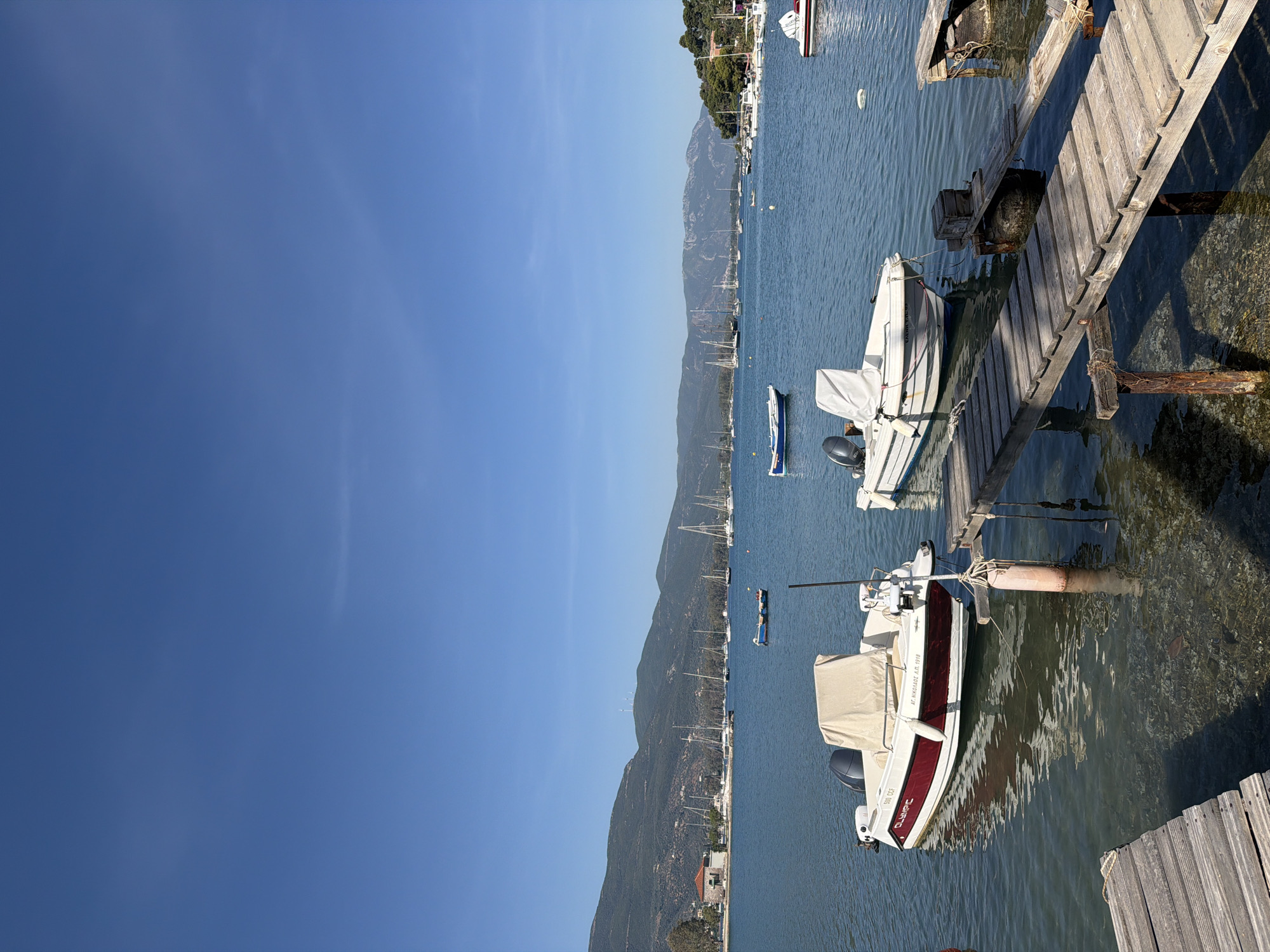 A tranquil daytime scene captures a vibrant Mediterranean coastal landscape. The image is dominated by a clear, deep blue sky and calm, azure waters. In the foreground, two weathered wooden docks extend into the water, with two small motorboats moored alongside. One boat, mostly white with a distinctive red stripe, bears the text "OLYMPIC" and "920 GG" and "Δ.Τ. ΜΥΚΟΝΟΥ Α.Α. 1918" on its hull. The other foreground boat, also white, has partially visible text "CARRY... L8F0" on its side. Both boats have outboard motors and are covered with light-colored tarpaulins.
Beyond the immediate docks, the water is dotted with numerous other boats, including several sailboats with their masts rising towards the sky, and smaller motorboats anchored or moored further out. The mid-ground reveals a long, verdant mountain range running horizontally, with scattered buildings and trees along its base, suggesting a coastal town or village. The sunlight is bright, creating clear reflections on the water and highlighting the textures of the wooden docks. The overall impression is one of peaceful maritime leisure under a cloudless sky in Poros, Greece.