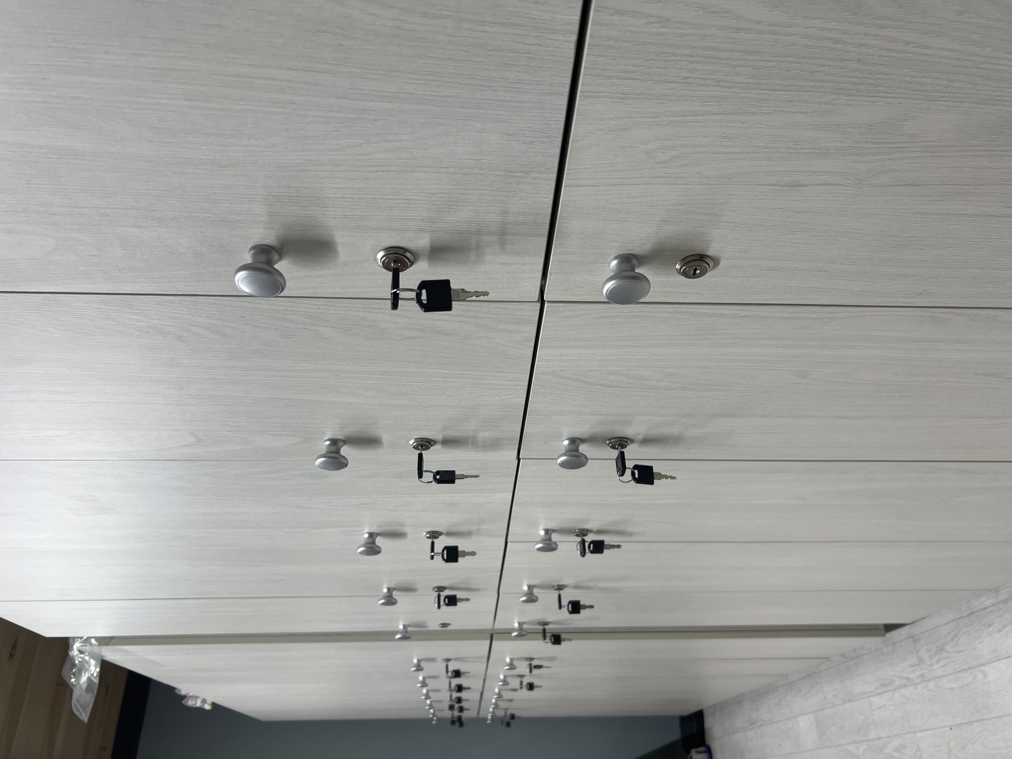 This image captures a series of modern, light grey or white laminate storage cabinets, likely drawers or lockers, arranged vertically in two columns. Each individual storage unit features a silver-colored, mushroom-shaped knob and a corresponding silver cam lock. Several black-headed keys are visibly inserted into the locks, suggesting they are either unlocked or recently secured.
The perspective is upward, creating a strong sense of depth and repetition as the cabinets extend far into the background, becoming smaller. The most striking feature is the highly reflective surface below the actual cabinets, creating an almost perfect mirror image of the entire setup. This reflection occupies the lower half of the photograph, effectively doubling the apparent height and number of storage units.
The laminate surfaces exhibit a subtle wood-grain texture. The lighting is bright and even, casting soft shadows from the knobs and keys, which adds to the three-dimensional quality of the hardware. In the bottom left corner of the main image, some wooden planks and clear plastic wrapping are faintly visible, and a darker area suggests the room's environment beyond the immediate cabinet structure and its reflections.
No specific activity is taking place; the scene depicts static furniture. The presence of keys in the locks implies the cabinets are in active use or prepared for access. The overall impression is one of cleanliness, order, and functionality, likely within an office, public facility, or storage area in Khabarovsk, Russia. There is no visible text.