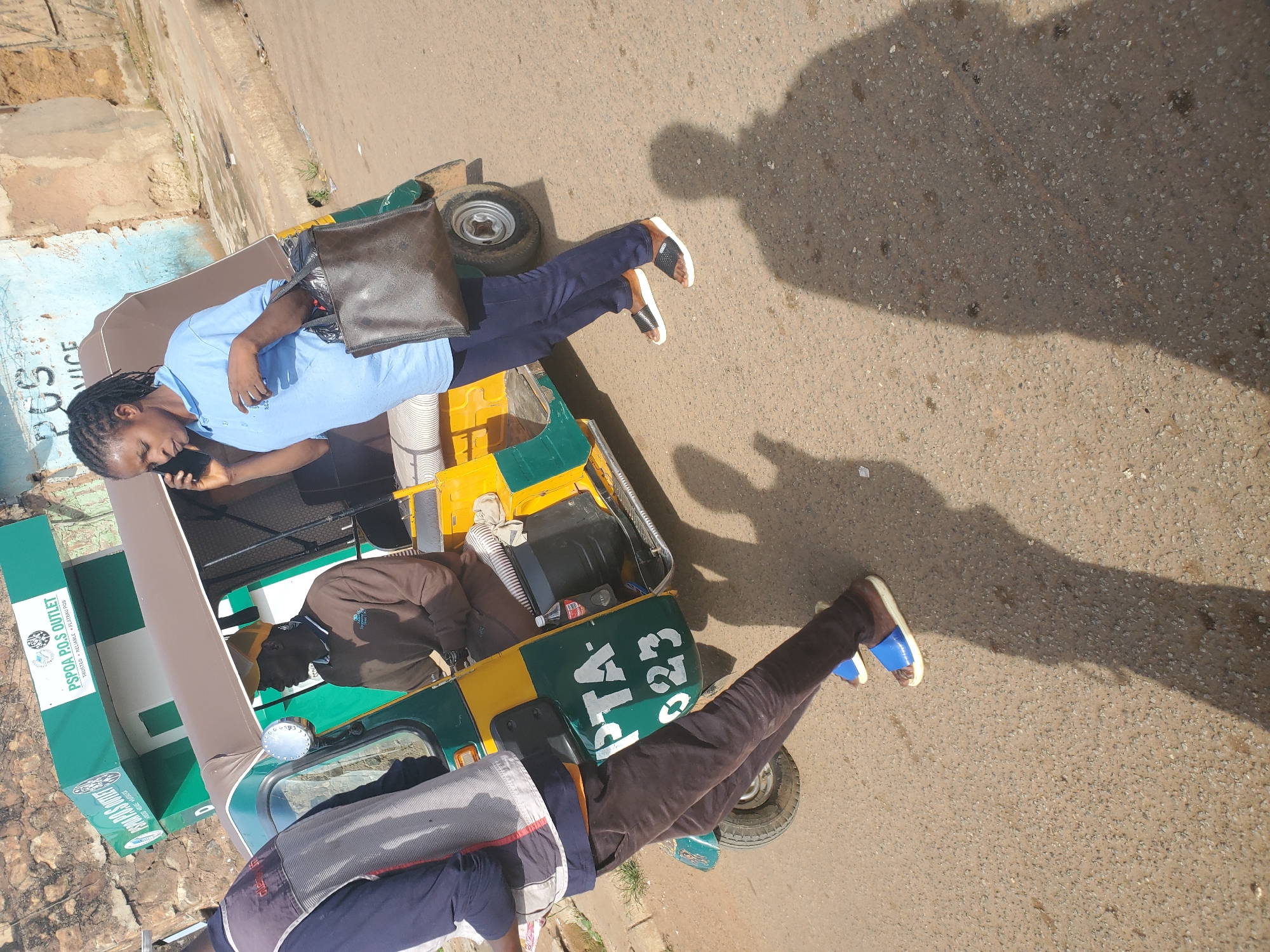 A medium shot shows a woman in a blue polo shirt and dark pants on a street in Bukuru, Nigeria. She is holding a dark brown purse and talking on a black smartphone. To her left is a yellow and green auto-rickshaw. The driver, wearing a brown long-sleeved shirt and a yellow cap, is visible in the driver's seat. To the left of the auto-rickshaw, a person in a dark blue jacket and grey vest stands facing away from the camera, their left leg partially obscuring the auto-rickshaw's rear tire. The auto-rickshaw has "PTA-023" painted in white on its green side panel. In the background, a green building with white lettering reads "PSPOA P.O.S OUTLET". The scene is illuminated by sunlight, casting long shadows on the asphalt road.