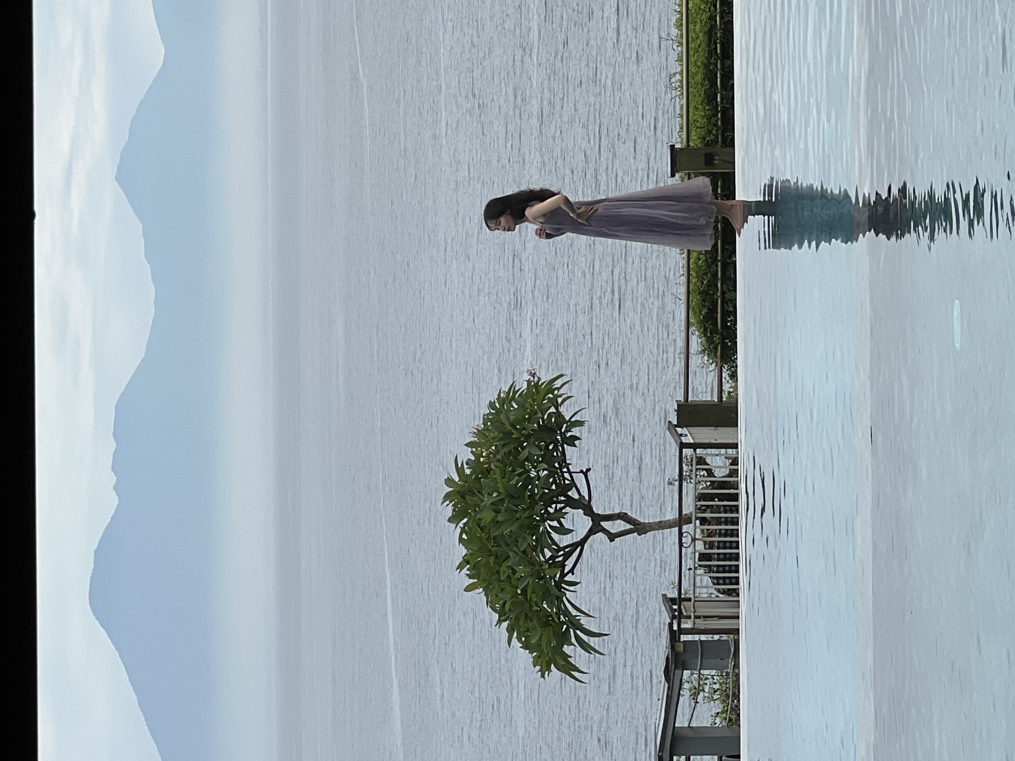 The image displays a tranquil outdoor scene, likely at a resort or hotel in Kuta Selatan, Indonesia. The perspective is from a patio area overlooking a vast body of water.
**Subjects:**
A young woman with long, dark hair, wearing a dark grey or muted purple sleeveless dress, stands near the edge of a foreground water feature, which appears to be a swimming pool or a still decorative pond. To her left and slightly closer to the viewer is a small, leafy tree with a distinctive, sprawling canopy, possibly a Frangipani. Behind the pool, a large body of water, likely the sea or a lake, extends to the horizon. In the distant background, gentle, hazy blue-grey mountains or hills are visible. The edge of a railing or fence is present to the right of the woman, suggesting a boundary of the patio area, with glimpses of outdoor furniture like chairs and tables.
**Activity or Event Taking Place:**
The woman is standing still, looking downwards into the foreground water. Her posture suggests a moment of contemplation or observation, possibly gazing at her reflection or the water's surface.
**Notable Details:**
The time of day appears to be daytime, with soft, diffused lighting, indicative of an overcast sky or a period when the sun is not directly overhead. The sky above the distant mountains is a pale, hazy blue, suggesting slightly cloudy or misty weather conditions. A prominent detail is the clear reflection of the woman and the tree in the still, clear water of the foreground pool. The larger body of water in the mid-ground exhibits subtle ripples. The scene evokes a sense of peace and relaxation, characteristic of a scenic coastal or lakeside retreat.
**Visible Text:**
There is no visible text in the image.