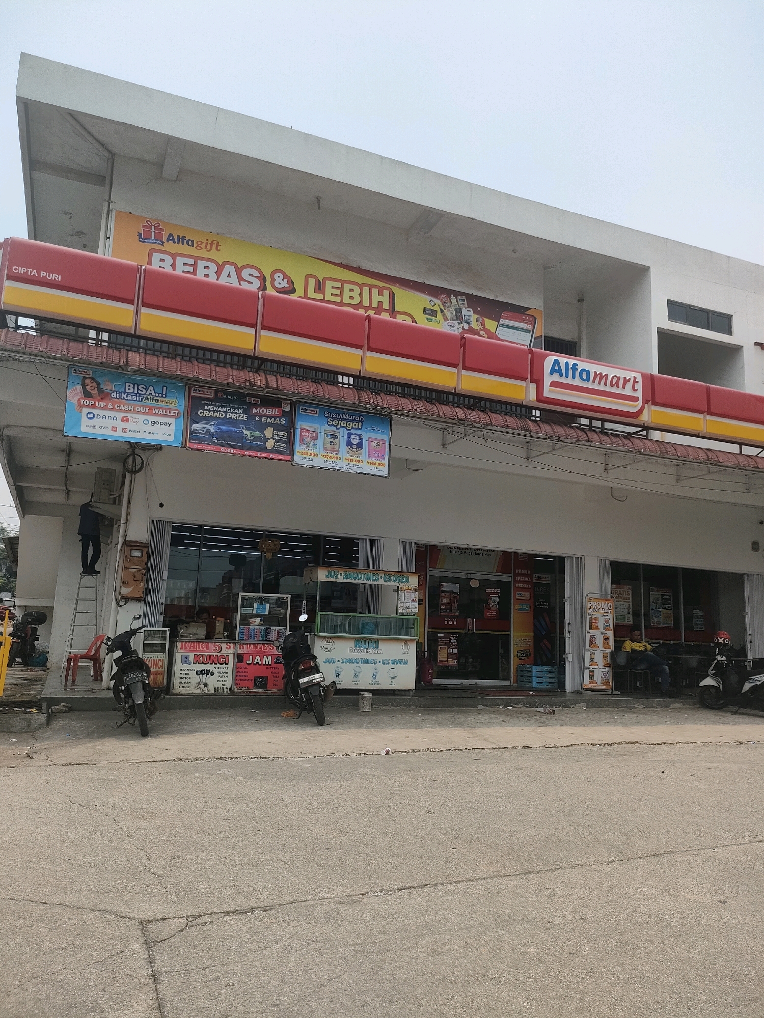The image shows a storefront of an Alfamart convenience store, a common chain in Indonesia, located on a street in Batam, Indonesia. The building is a single-story structure with a white facade and a prominent red and yellow awning above the entrance. Several advertisements are displayed on the facade, including one for "Alfamart" and another promoting "BEBAS & LEBIH".
In front of the store, there are several motorcycles parked. The street in front of the store is paved with asphalt and shows signs of wear. A person is seen on a ladder, possibly performing maintenance work on the building or its signage. Inside the store, shelves stocked with products are visible through the glass entrance.
To the left of the entrance, there is a stand with various promotional materials and signs. To the right, a few people are seated, seemingly taking a break. The overall scene depicts a typical commercial street in an Indonesian city.