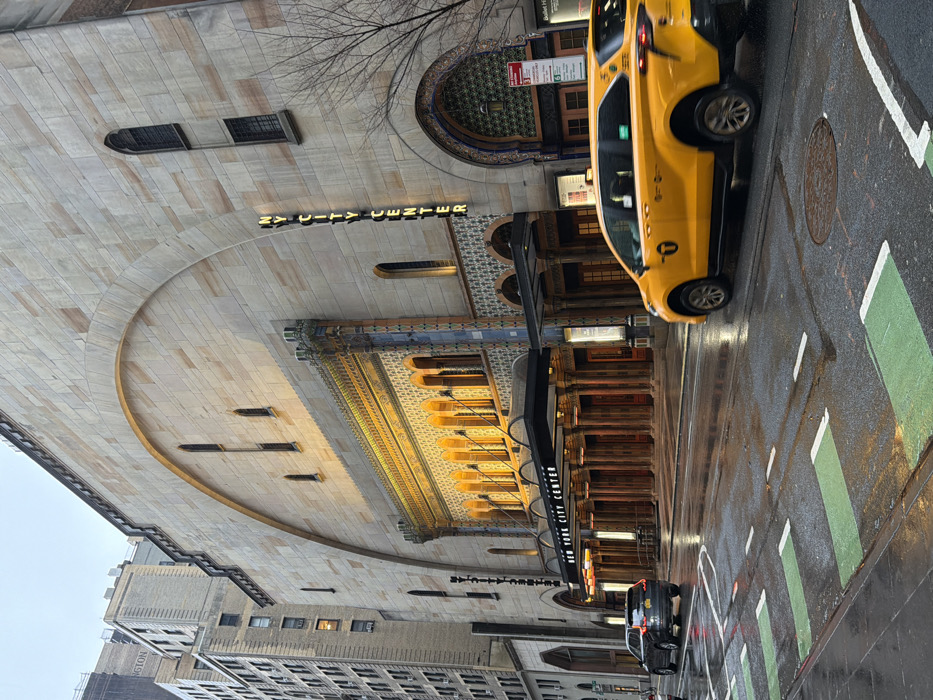 The image depicts a street scene in front of the New York City Center, located at 131 W 55th Street in New York, United States of America.
The New York City Center building dominates the frame, showcasing its distinctive large, light-colored stone facade featuring a prominent upper arch. The lower section of the building includes intricate architectural details, such as multiple arched windows and decorative mosaic-like patterns, illuminated by warm yellow lights. A large black awning extends over the main entrance, displaying "NEW YORK CITY CENTER" in white lettering. A vertical sign spelling "NY CITY CENTER" is affixed to the right side of the building's facade. To the left, a taller, multi-story building with numerous windows is partially visible.
The street in the foreground is wet asphalt, indicating recent precipitation. It features painted green sections with white dashes, consistent with bicycle lane or pedestrian crossing markings. A circular manhole cover is visible on the street surface. Two vehicles are in motion: a dark gray SUV, likely a Volvo, is positioned in the left lane, moving away from the viewer with its rear lights illuminated. A yellow New York City taxi minivan, identifiable by its color, "T" medallion logo, and vehicle type (likely a Toyota Sienna), is in the right lane, moving from right to left across the frame, with slight motion blur evident. Bare tree branches are visible on the right side of the frame, suggesting a leafless season. The overall lighting suggests overcast conditions or early twilight. No discernible individuals are clearly visible.