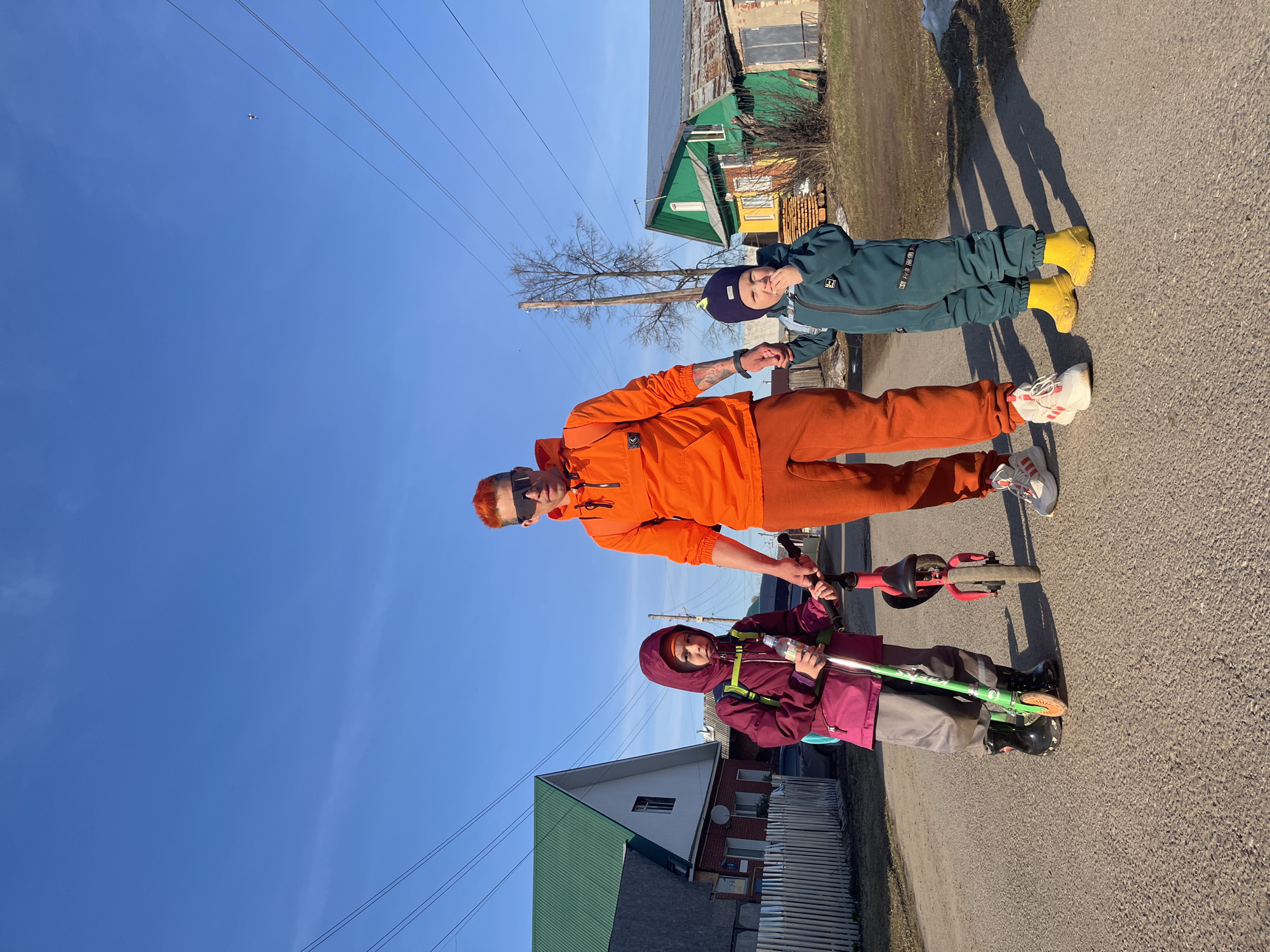 The image shows a group of three individuals standing on a paved surface, likely a street or pathway, in what appears to be the city of Chaikovsky, Russia, based on visual cues like architecture and signage. The sky is clear and blue, suggesting a sunny day.
A person in the middle wears a bright orange hooded jacket and matching pants, along with white sneakers. They are holding hands with the other two. One of them is a child clad in a dark blue/green jumpsuit and yellow boots, also wearing a dark blue hat. The other is a child in a reddish-purple hooded jacket and light grey pants, holding a green scooter. There is another scooter in the hands of the person in orange.
In the background, there are houses and buildings typical of a rural or suburban area. Utility poles and wires are visible against the clear sky. There is a mix of green and brown vegetation present. Shadows of the individuals are cast on the paved surface.