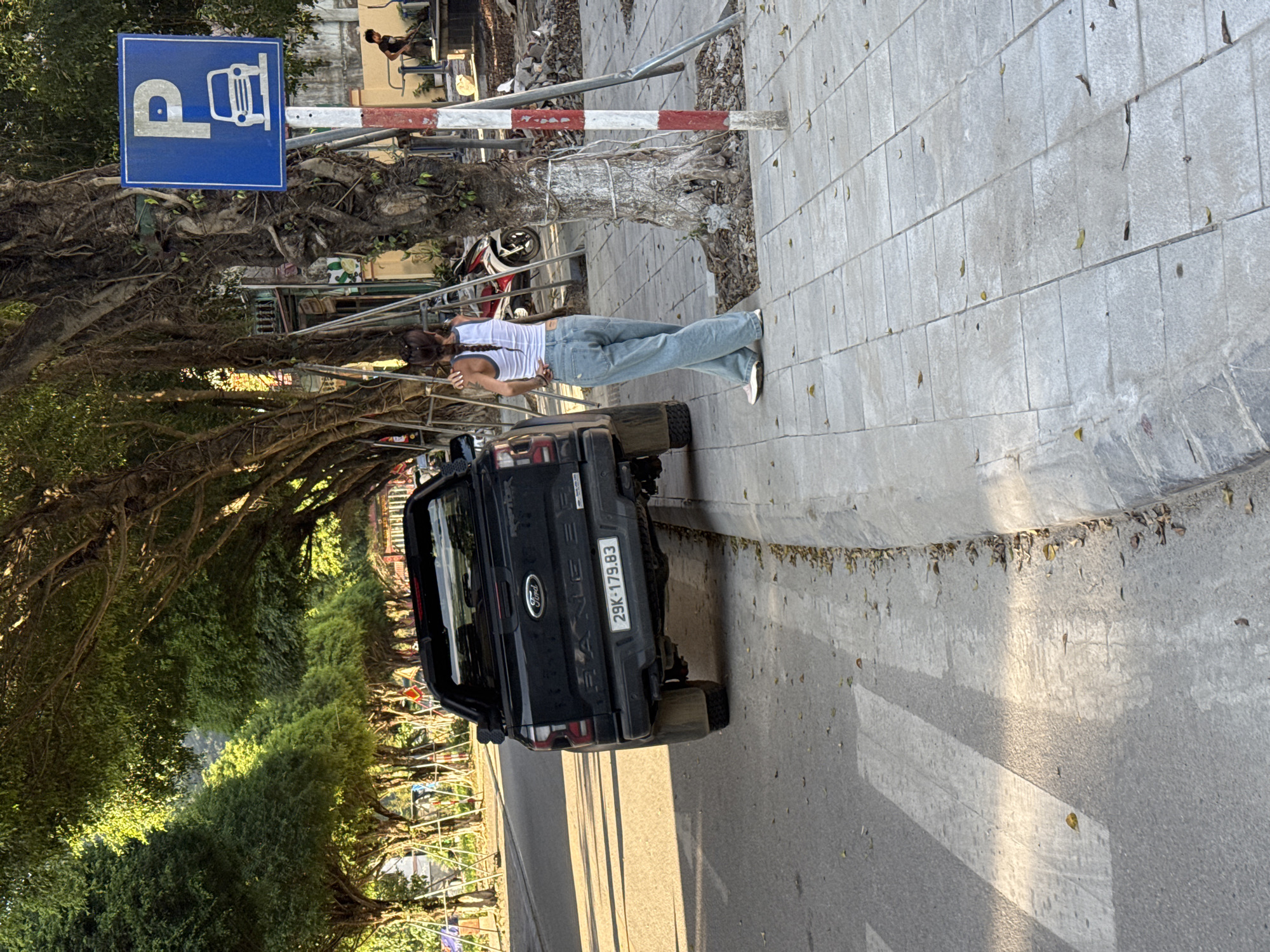A long shot captures a black Ford Ranger truck parked in Lao Cai Ward, Vietnam. The vehicle, bearing license plate 29K-179.83, is situated on the side of a road lined with lush green trees. A woman dressed in a white tank top and light blue jeans stands near the truck. Adjacent to the truck and the woman, a sidewalk paved with gray stones is visible. A blue parking sign with a white "P" and a bus symbol stands nearby, mounted on a metal frame with red and white stripes.