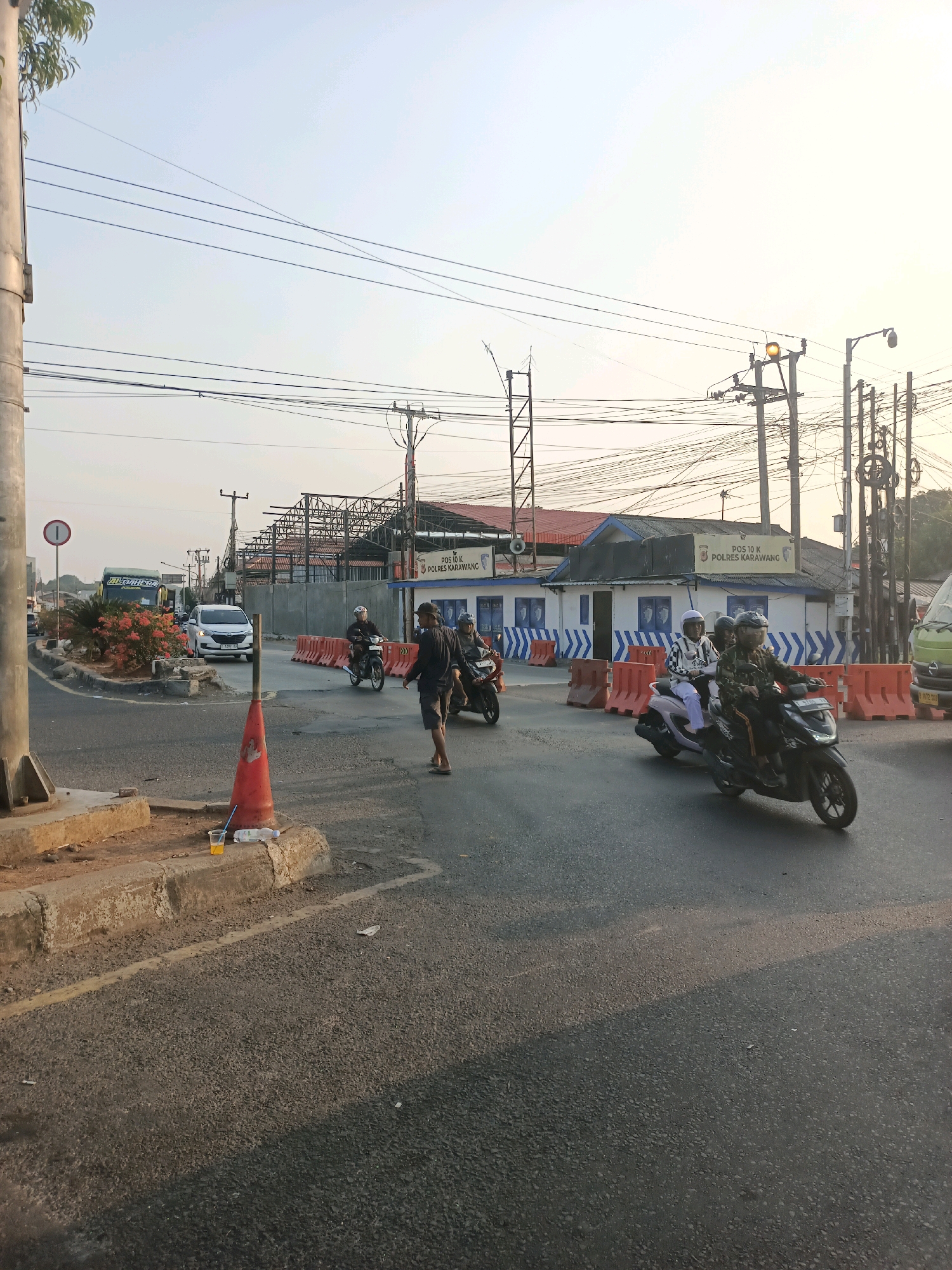 The image depicts a street scene in Karawang, Indonesia, likely near a police post. In the foreground, a concrete curb is visible with an orange traffic cone and a discarded plastic bottle and cup with a straw. The street is paved with asphalt, and several motorcycles are in motion. One motorcycle carries two individuals wearing helmets. Another motorcycle, ridden by a single person in a helmet, is in the process of passing. Further down the road, another motorcycle with two occupants, both wearing helmets, is visible. A white car is also present, along with a large green bus.
In the background, a low-rise building with blue and white detailing is identifiable as "POS 10 K POLRES KARAWANG," indicating it is a police post in Karawang. Orange traffic barriers are strategically placed along the road. Overhead, a complex network of electrical wires and poles dominates the sky, suggesting an urban or semi-urban environment. The sky is clear and bright, indicating daytime, possibly late afternoon given the lighting and shadows. Some trees are visible on the left side of the image, and the skeletal structure of a building under construction or renovation is present in the distance. The overall scene conveys a sense of everyday traffic and activity at a roadside location with a police presence.