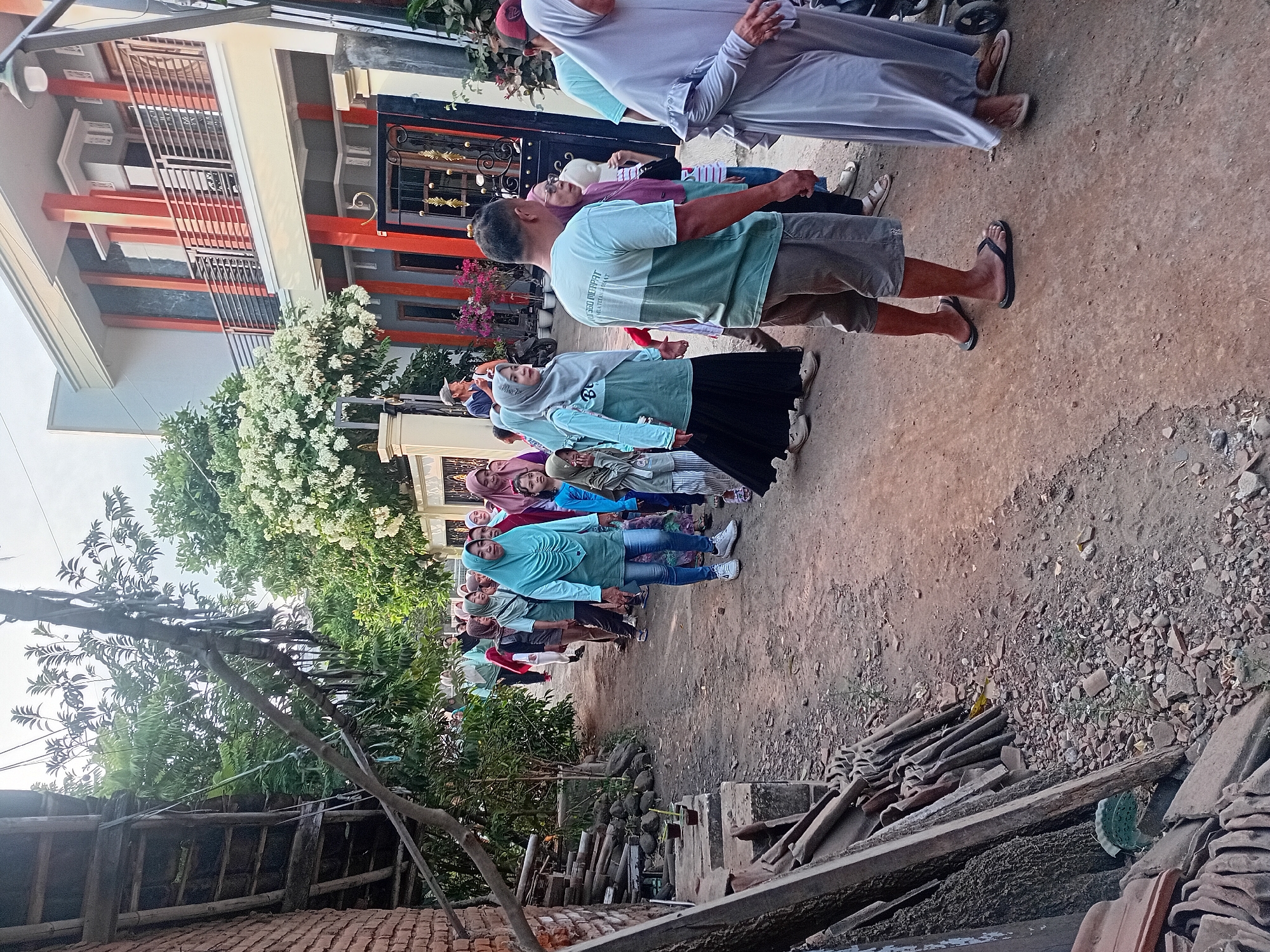 In Besito, Indonesia, a group of people, predominantly women and girls wearing hijabs and modest clothing, are walking in a line down a dirt path lined with trees and buildings. A man in a green and grey t-shirt and shorts is walking towards the camera in the foreground. To the left of the path, there is a brick wall and a stack of wood and tiles. A modern house with orange accents is visible in the background on the right. The sky is overcast, suggesting it might be late afternoon or early morning.