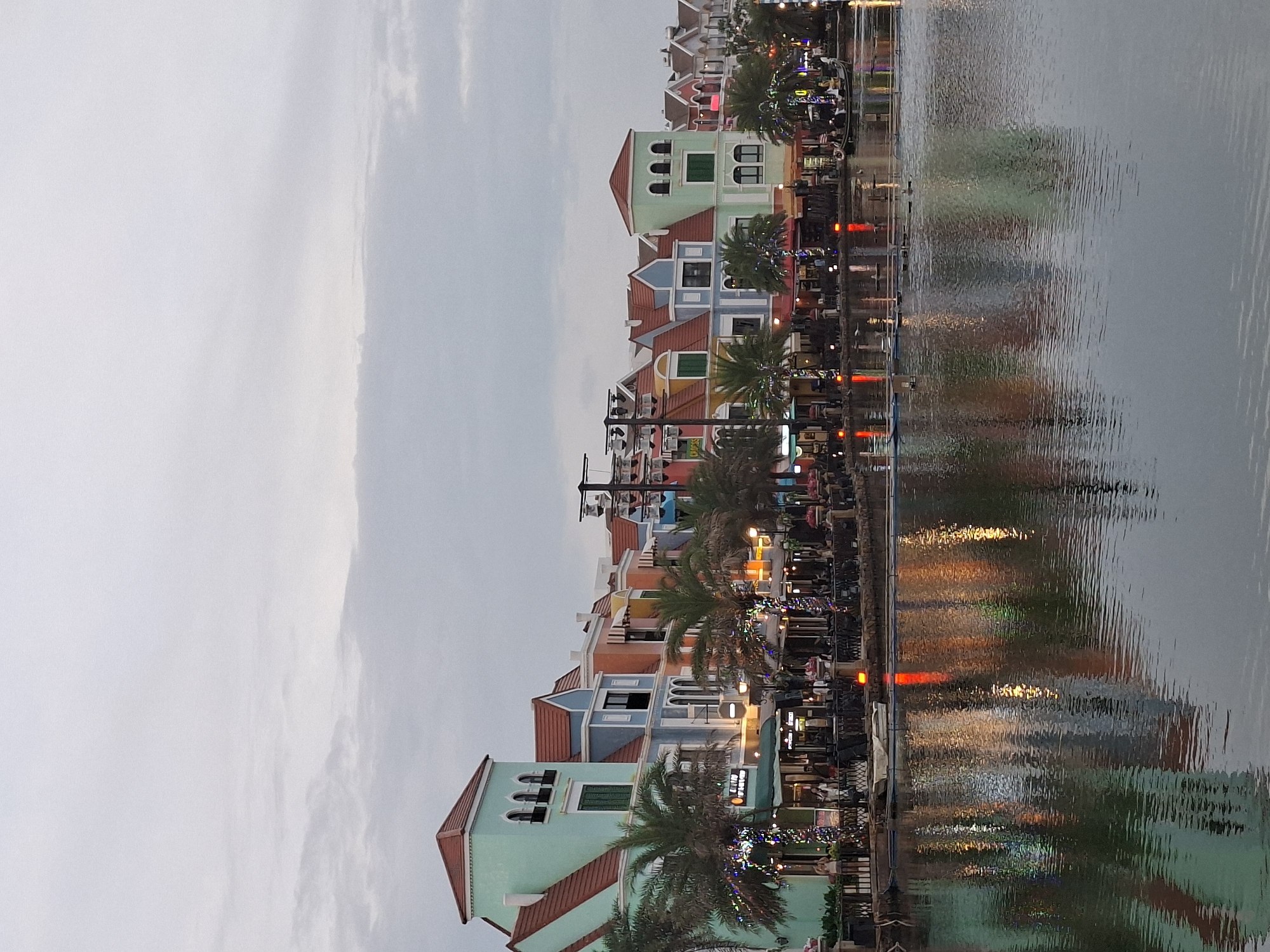 This image depicts a waterfront scene in the city of Phú Quốc, Vietnam, likely at a commercial or entertainment area. A row of colorful buildings, resembling European architecture with pastel facades and red tiled roofs, lines the waterfront. These buildings feature arched windows and balconies, with palm trees in front of them. String lights are visible, indicating an evening setting or festive decoration.
The buildings house what appear to be restaurants or shops, with awnings and outdoor seating areas. Several people are visible on the sidewalk in front of the establishments, some standing and others seated. Lights from within the buildings and from the decorative string lights reflect on the water.
A calm body of water, likely a canal or a man-made lake, separates the viewer from the buildings. The water's surface is slightly rippled, creating distorted reflections of the architecture and lights. A small boat is present on the water, near the shore.
The sky is overcast and muted, suggesting either early morning or late evening. In the background, more buildings and structures, including what appear to be utility poles with lights, can be seen. The overall impression is of a picturesque and lively waterfront district.