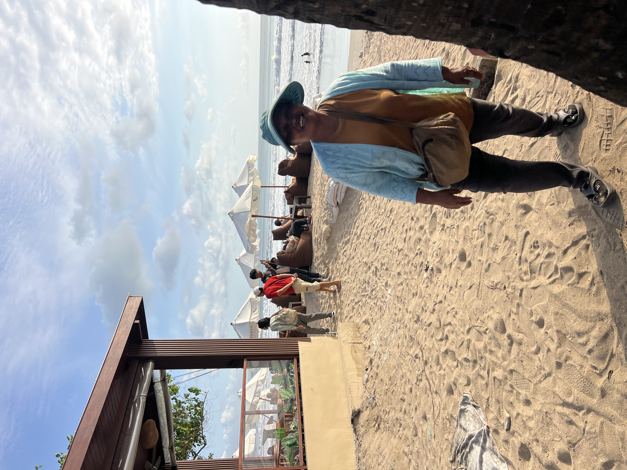 This is a medium shot photograph taken on a sandy beach in Kuta, Indonesia. A woman wearing a blue cardigan, an orange shirt, dark pants, and a blue hat stands in the foreground, smiling at the camera. She has a brown messenger bag slung across her body. Behind her, the beach extends towards the ocean, with several white umbrellas and beanbag chairs set up. In the background, people can be seen walking and relaxing, and two individuals are in the water. A structure with a brown roof and wooden pillars is visible on the left side of the image. The sky is partly cloudy, and the sun appears to be setting, casting a warm light over the scene.