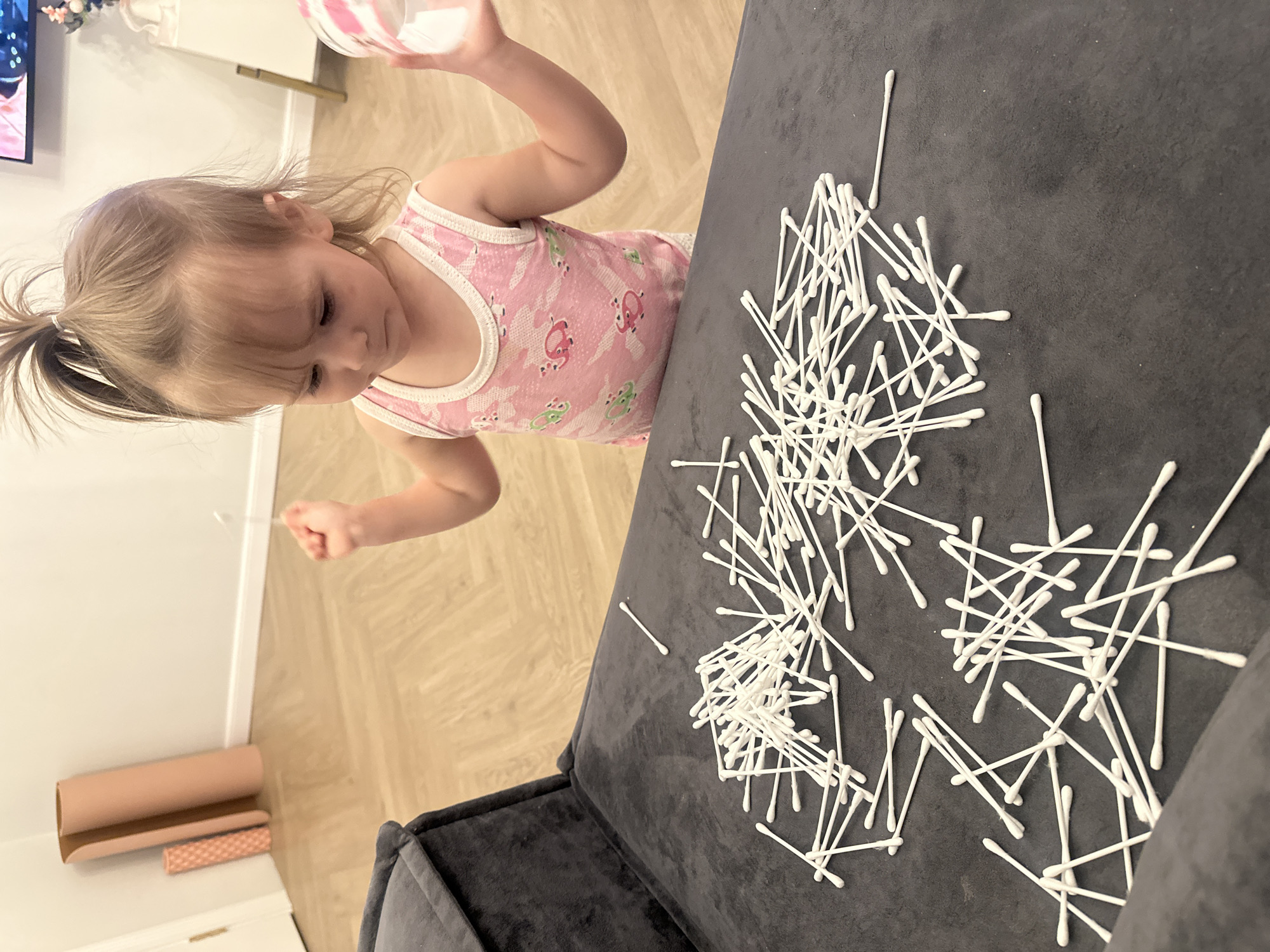 A young child with blonde hair in a ponytail, wearing a pink tank top with an animal print, stands near a dark gray upholstered sofa. The child holds a clear plastic container in one hand and appears to be looking at or interacting with a large pile of white cotton swabs spread across the sofa's surface. The setting appears to be indoors with light-colored wooden flooring. To the left, there is a rolled-up piece of paper or cardboard and a small patterned object on a white cabinet. A television is visible in the far upper left corner. The location is not possible to determine based solely on the image.