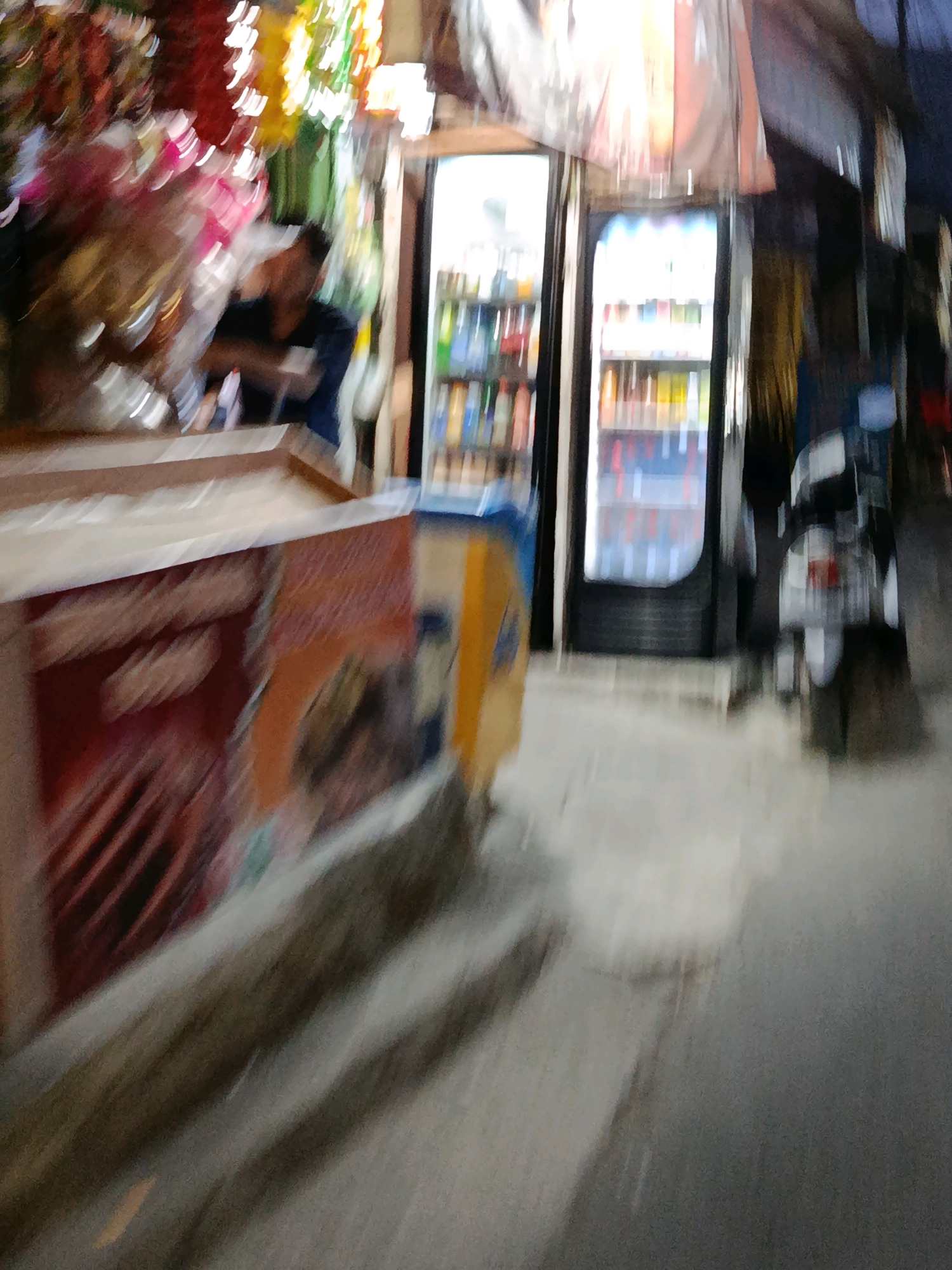 The image depicts a blurry scene in front of a convenience store in the city of Bekasi, Indonesia. A person, whose face is obscured by blur, is visible inside the store, standing near a display of colorful items. Outside the store, two glass-door refrigerators are stocked with beverages. To the right of the refrigerators, a motorcycle is parked. The foreground shows a concrete walkway, suggesting an urban setting. The lighting indicates it may be evening or nighttime.