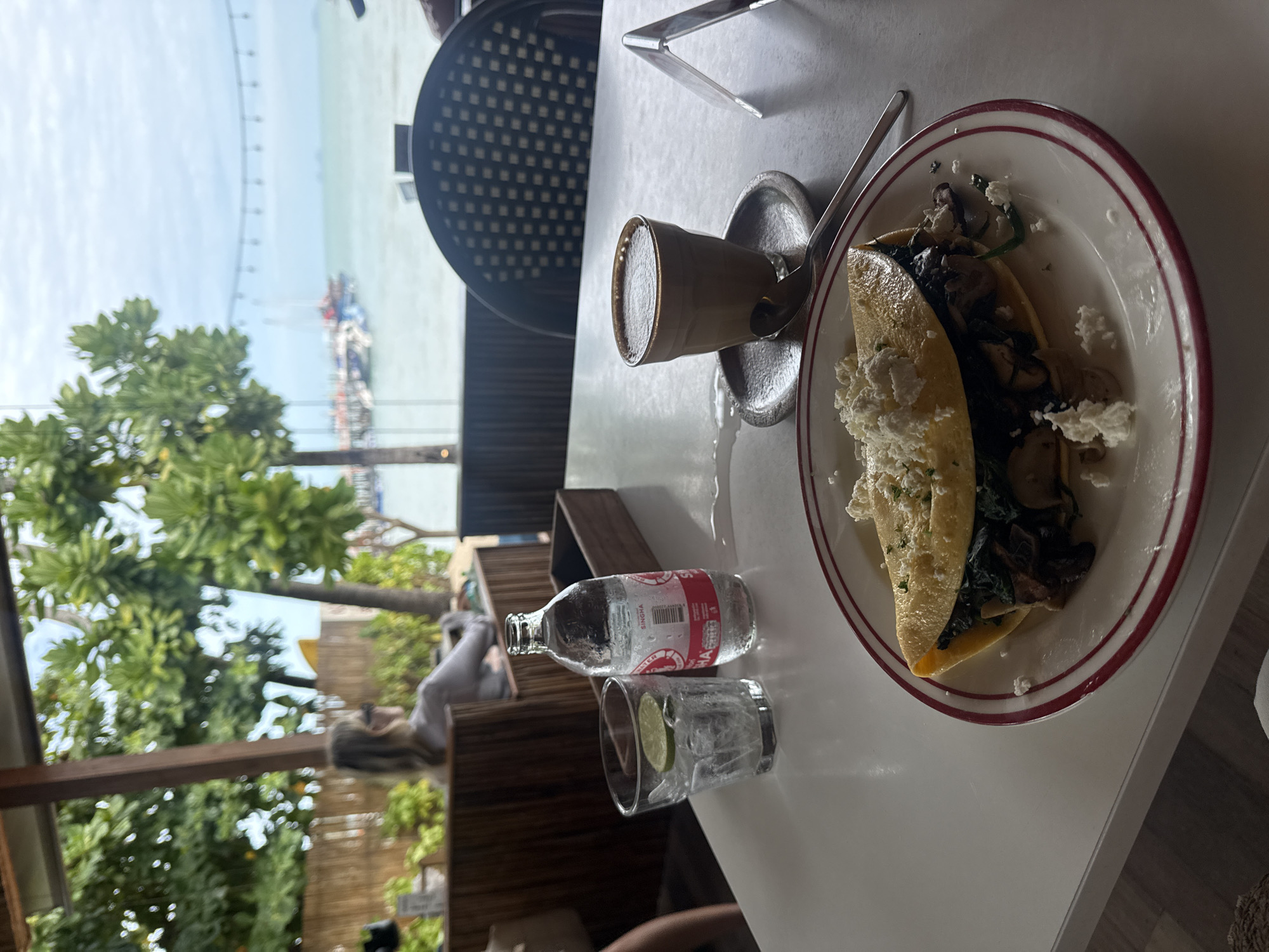 In Ko Samui District, Thailand, a table at a seaside restaurant displays a meal. In the foreground, a white plate with a red rim holds a folded omelet filled with spinach and mushrooms, topped with crumbled white cheese and herbs. To the right, a small cup of coffee sits on a saucer, with a spoon resting beside it. Further left, a glass filled with ice and a lime wedge sits next to a bottle of Singha water. In the background, a woman with blonde hair, wearing sunglasses and a light-colored top, is seated at a wooden booth, looking out towards the ocean. The restaurant appears to be open-air, with lush green trees and a view of boats docked in the water under a cloudy sky.