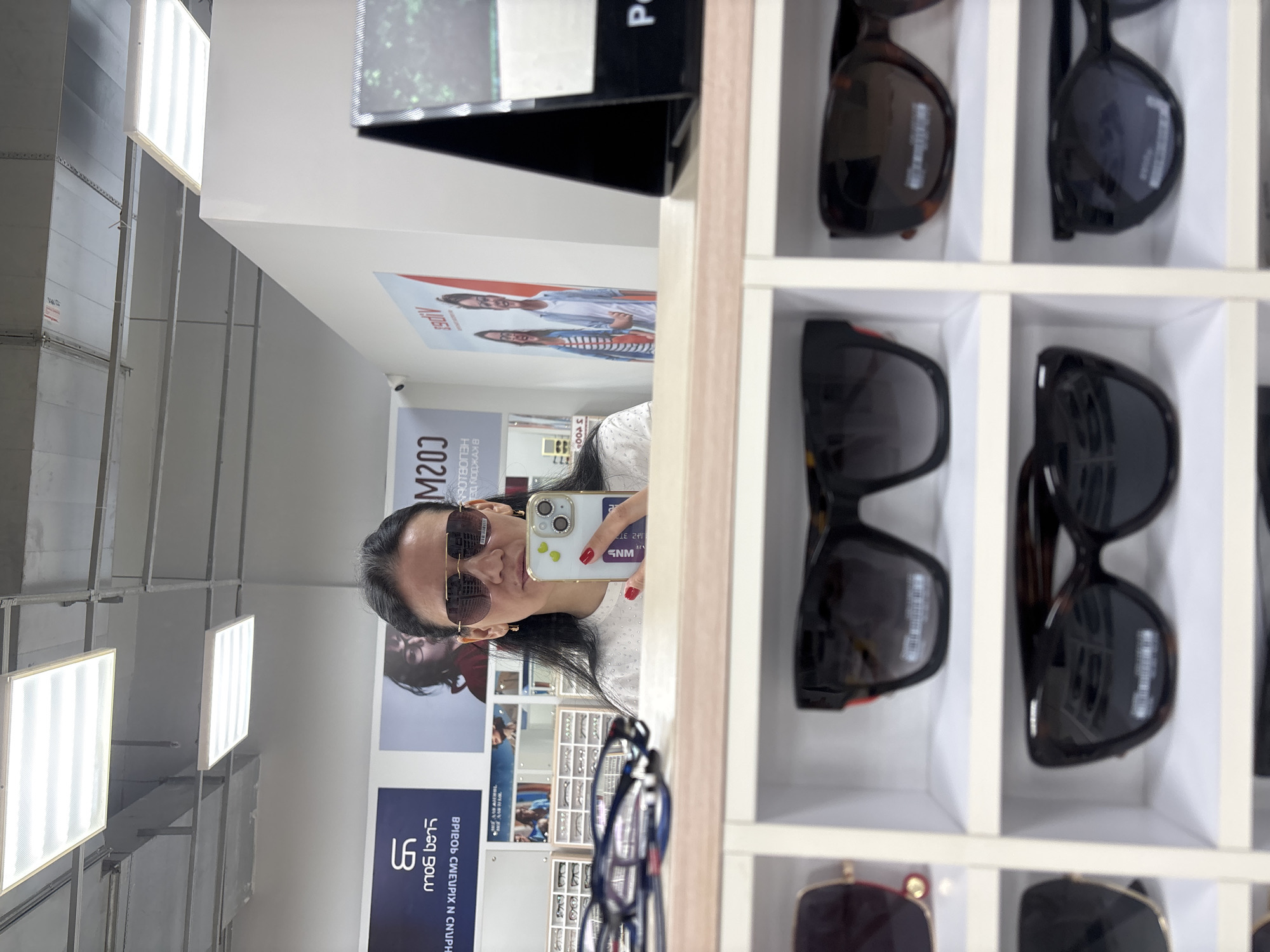 The image depicts a woman in Chaikovsky, Russia taking a selfie in a store displaying eyewear. The location is evident through the Russian text on promotional materials visible in the background. The woman is holding her smartphone with a decorated case and wearing sunglasses, seemingly trying them on in front of a mirror. The store showcases numerous pairs of sunglasses organized in white compartments, revealing a diverse selection. The ceiling is visible, featuring rows of fluorescent lighting fixtures.