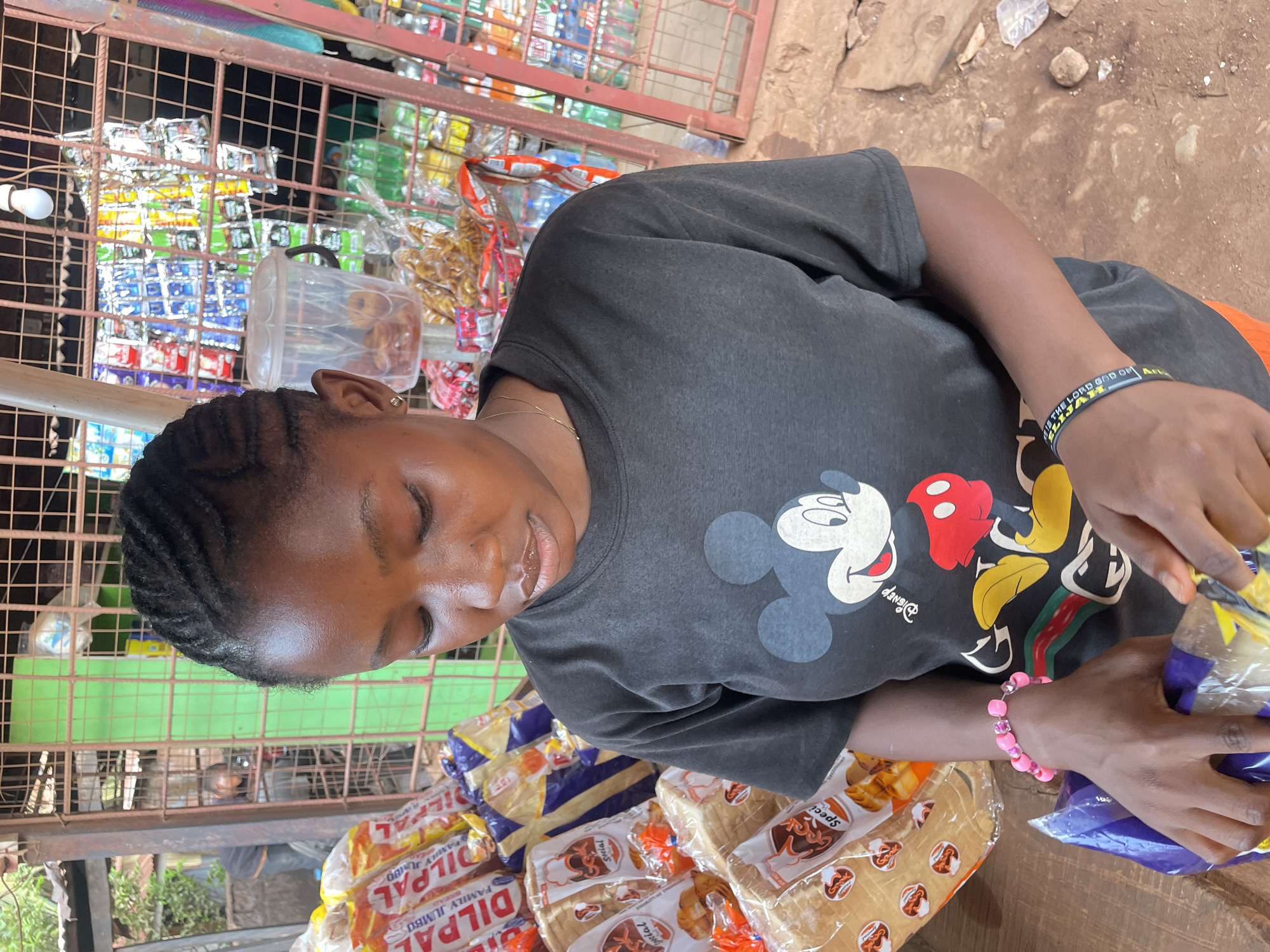 A woman with braided hair wearing a Mickey Mouse t-shirt is at a stand in Otukpa, Nigeria. She has a pink beaded bracelet on her right wrist and a black and yellow wristband on her left that reads “Elijah is the Lord God of”. She is bagging bread with labels “Special” and “Dilpa Family Jumbo” on a wooden table. Behind her is a metal grid displaying various items.