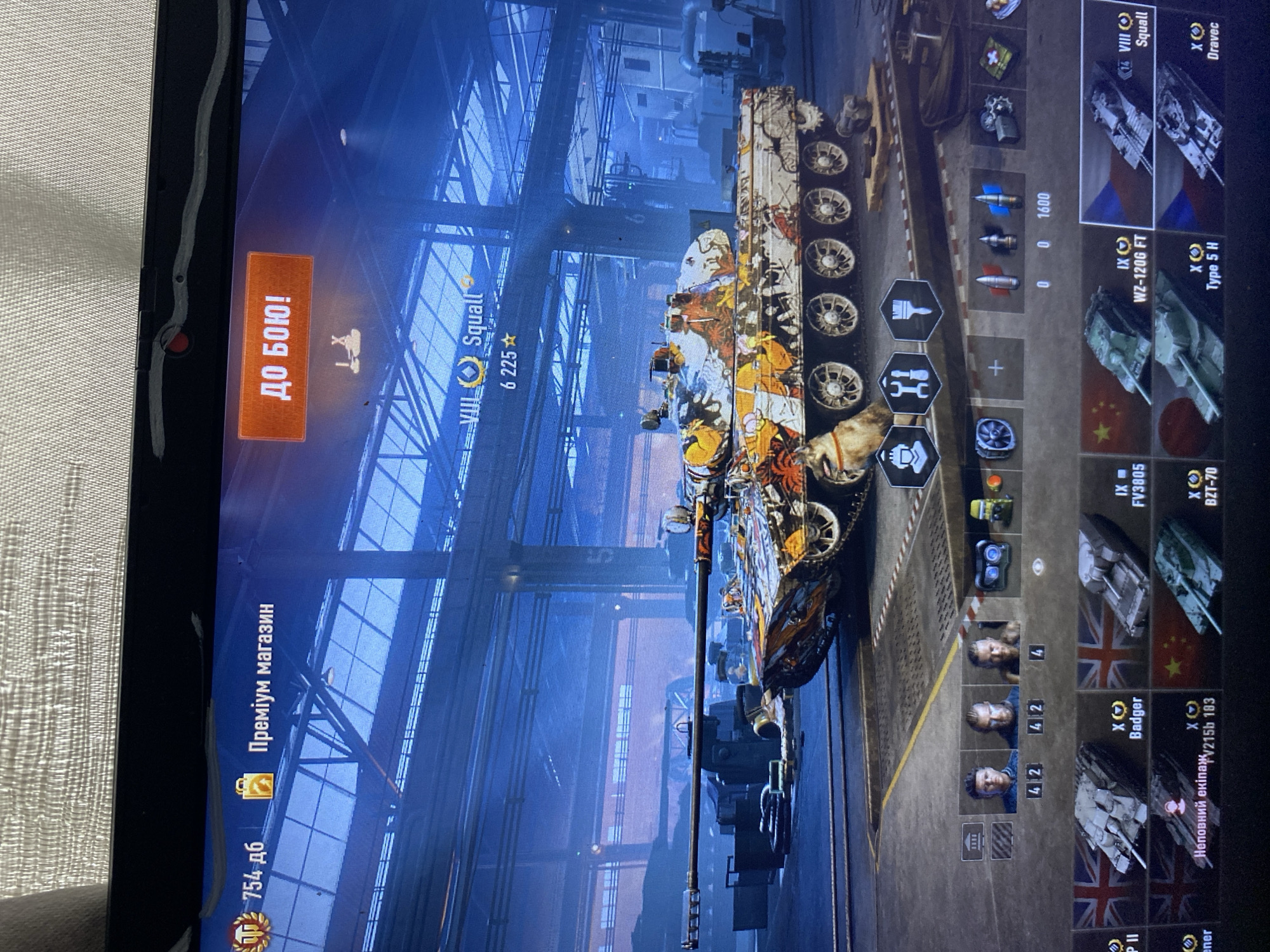 The image shows a screenshot of a video game, likely World of Tanks, displayed on a laptop screen. The game interface is visible, with elements like game currency (754 дб), a "Premium shop" button (Преміум магазин), and a "To battle!" button (ДО БОЮ!). A virtual tank with a distinctive orange and white camouflage pattern is prominently displayed in the center of the screen. Below the tank, there are icons representing various game features and modules, as well as a selection of tanks with their national flags. Crew members are also visible in the interface. The background of the game depicts an industrial hangar setting with a blue-toned lighting scheme. The laptop itself has a textured surface on the top edge and a webcam lens. There is no visible information within the image that provides any context or indication of the location being the city of Szczecin, Poland.
