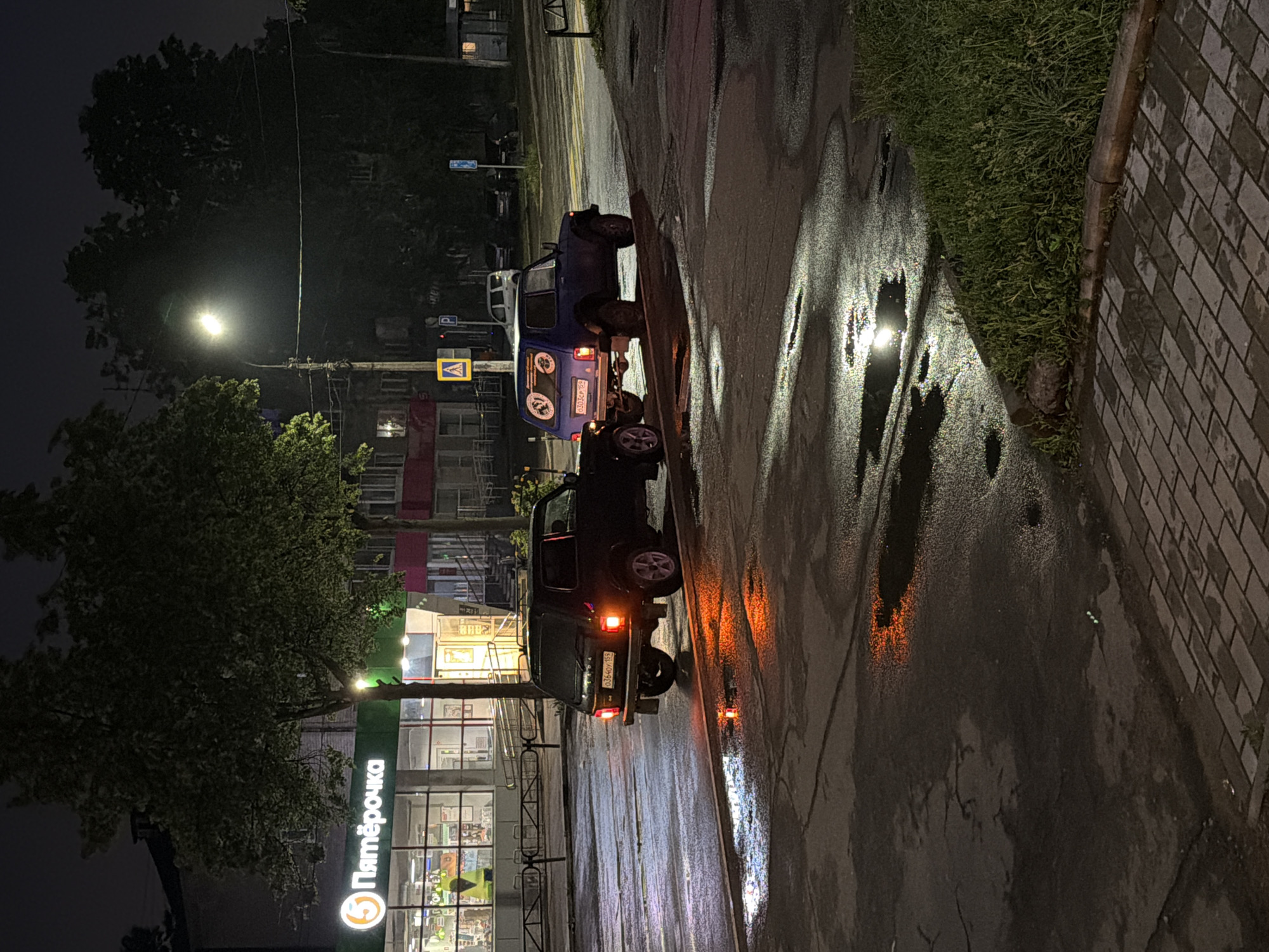The image depicts a nighttime street scene in Chaikovsky, Russia, characterized by a wet asphalt road reflecting various lights, suggesting recent rainfall. Two off-road vehicles are parked on the side of the street. The car further down the road is a blue SUV with a spare tire mounted on the rear and a round, possibly club-related, decal on its back window. Its Russian license plate, "0102PP 159", is visible. Closer to the foreground, a dark-colored SUV is parked, also featuring a rear-mounted spare tire and illuminated red/orange taillights. Its license plate reads "O304PY 159".
On the left side of the street, a large tree partially obscures a building with extensive glass windows, which houses a brightly lit "Пятёрочка" (Pyaterochka) supermarket, a prominent Russian retail chain. A street light casts a strong glow in the upper left, contributing to the reflections on the wet ground. To the right, a paved sidewalk with grey and light-colored bricks runs alongside a patch of grass. Street signs visible include a yellow triangular pedestrian crossing sign and a blue parking sign. The scene is devoid of people, conveying a quiet atmosphere.