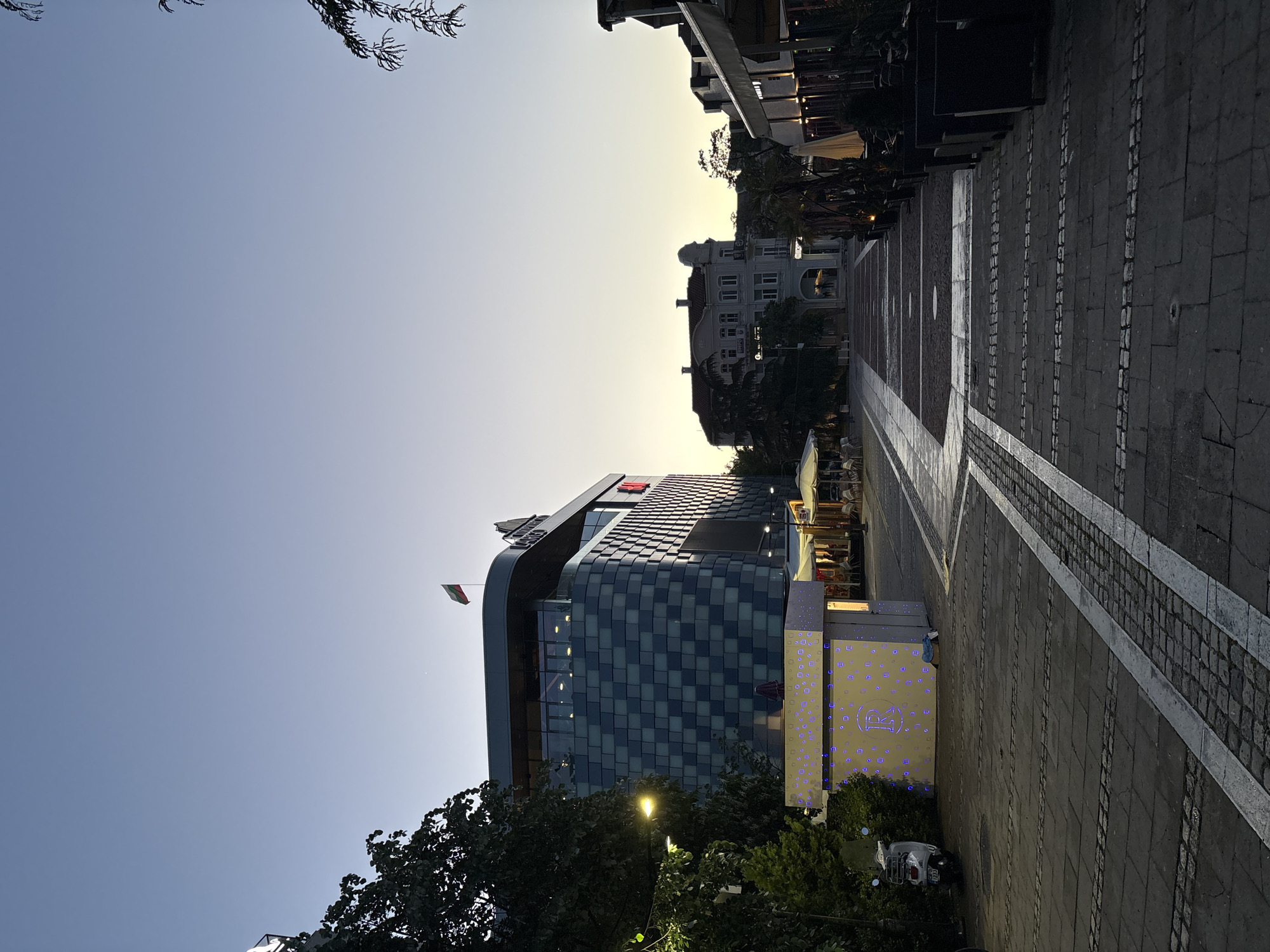 The image shows a street scene in Blagoevgrad, Bulgaria. The view is dominated by a modern building with a checkered glass facade, identified as the "LARGO MALL Blagoevgrad," with a Bulgarian flag flying from its roof. A pedestrian walkway with a patterned stone surface leads away from the building. Trees partially obscure the lower portion of the building and sidewalk. Streetlights are visible. In the distance, another building is seen along the walkway. The sky transitions from blue to a yellowish hue, suggesting dawn or dusk. A scooter is parked near the mall entrance.