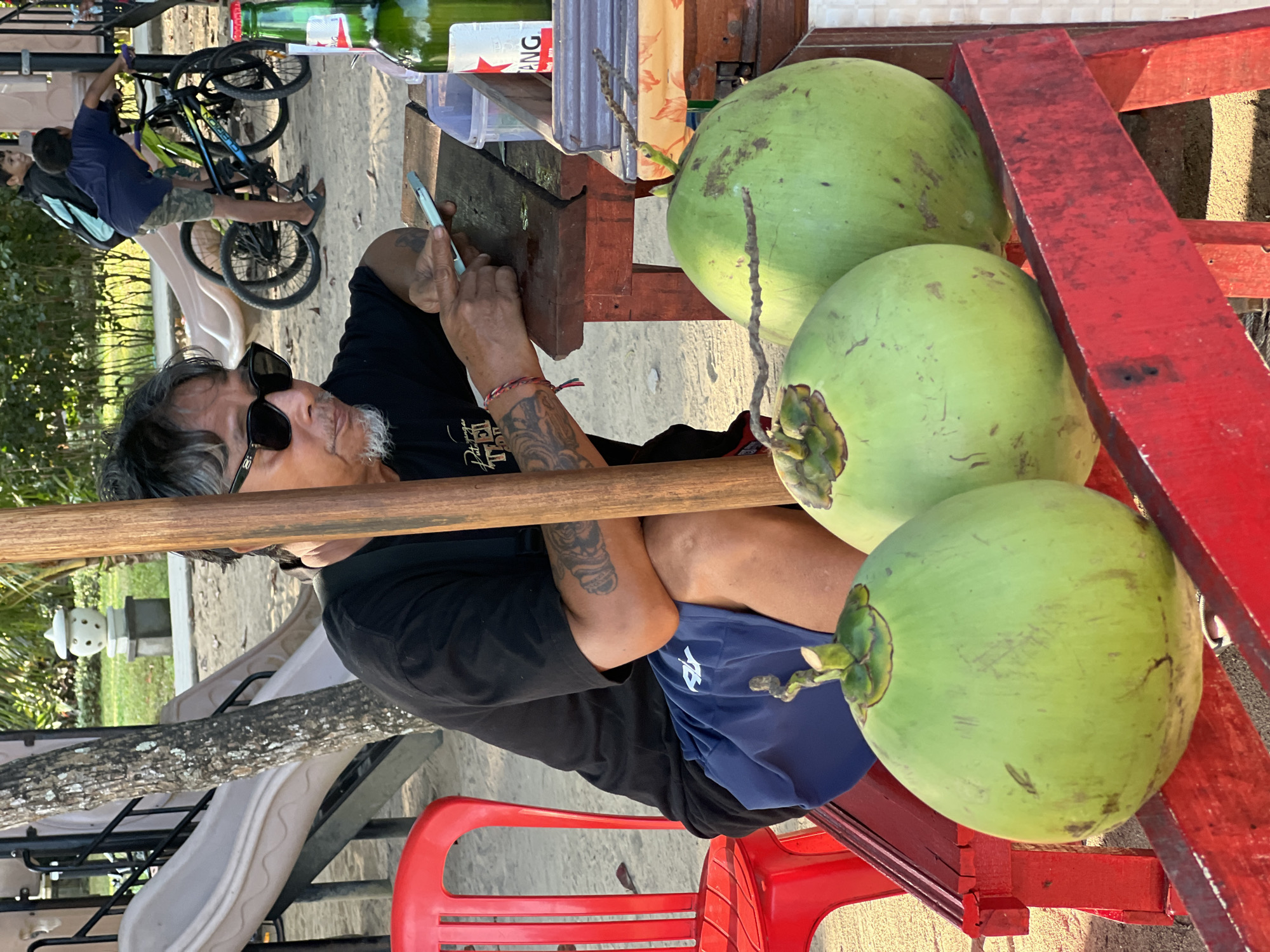 The image depicts a street scene in Denpasar, Indonesia, with a focus on a man selling coconuts. The man, wearing a black t-shirt and blue shorts, is seated behind a makeshift stall, holding a mobile phone in his hands. He has tattoos on his arms and is wearing sunglasses. In front of him are three green coconuts displayed on a red wooden stand. To his left, a red plastic chair is visible. In the background, children are playing near a playground with a slide, and bicycles are parked nearby. A green bottle of Bintang beer, a popular Indonesian brand, sits on the stall. The ground appears to be sandy, consistent with a beachside location or a park. The overall atmosphere suggests a casual, outdoor setting.