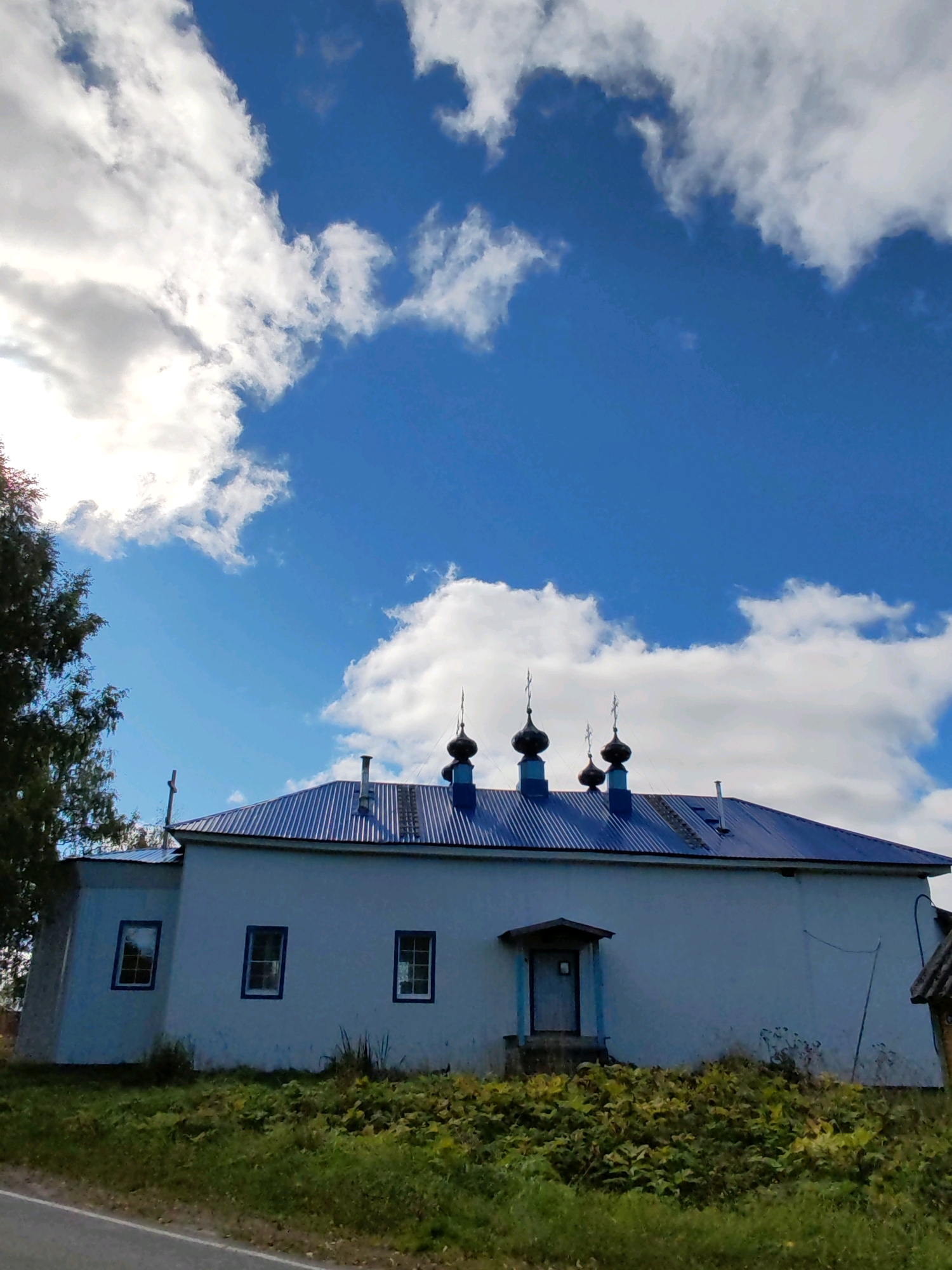 The image shows a single-story building with a blue, corrugated metal roof, situated in a rural area. The building has white walls and several rectangular windows with blue frames. There are multiple onion domes with crosses on top of the roof, indicating it is likely a church or chapel. A small porch with a door is visible on the right side of the building. The foreground is covered in green grass and vegetation, with a road or path running along the bottom edge of the frame. In the background, a tree is visible on the left, and the sky is a bright blue with scattered white clouds. The location is identified as Чухлэм, Russia.